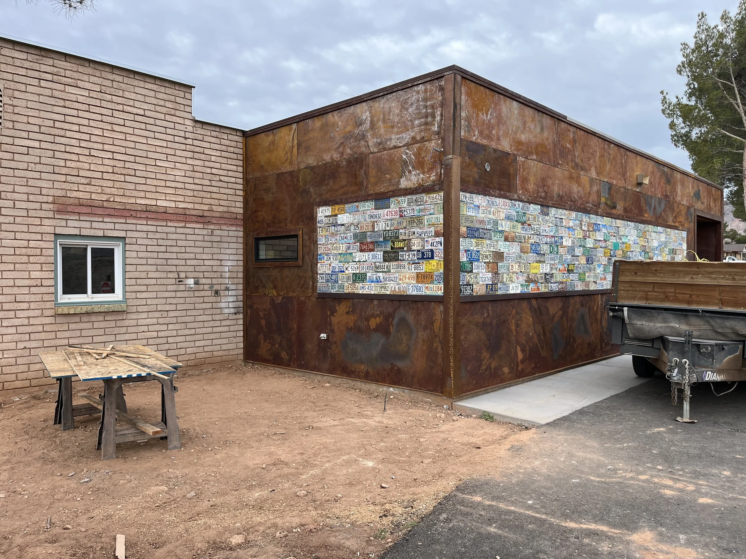 A building with a brick exterior on the left and a rust-colored metal exterior on the right, featuring a large window decorated with multiple license plates, under a cloudy sky. There is a small window on the brick section and construction work tools