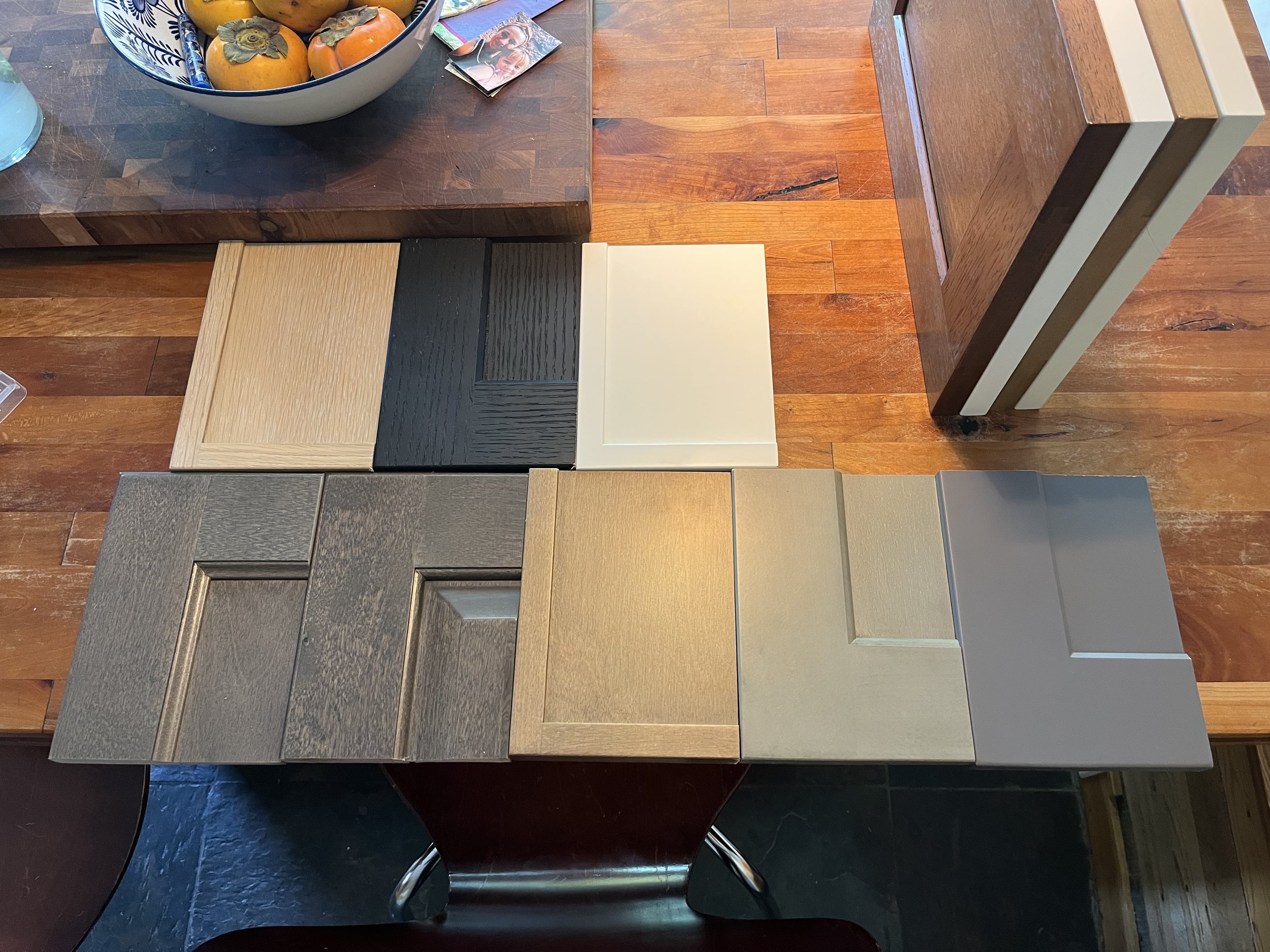 A wooden table displays sample square tiles in various shades of gray, brown, and beige, alongside a round ceramic bowl of persimmons and some small photographs.