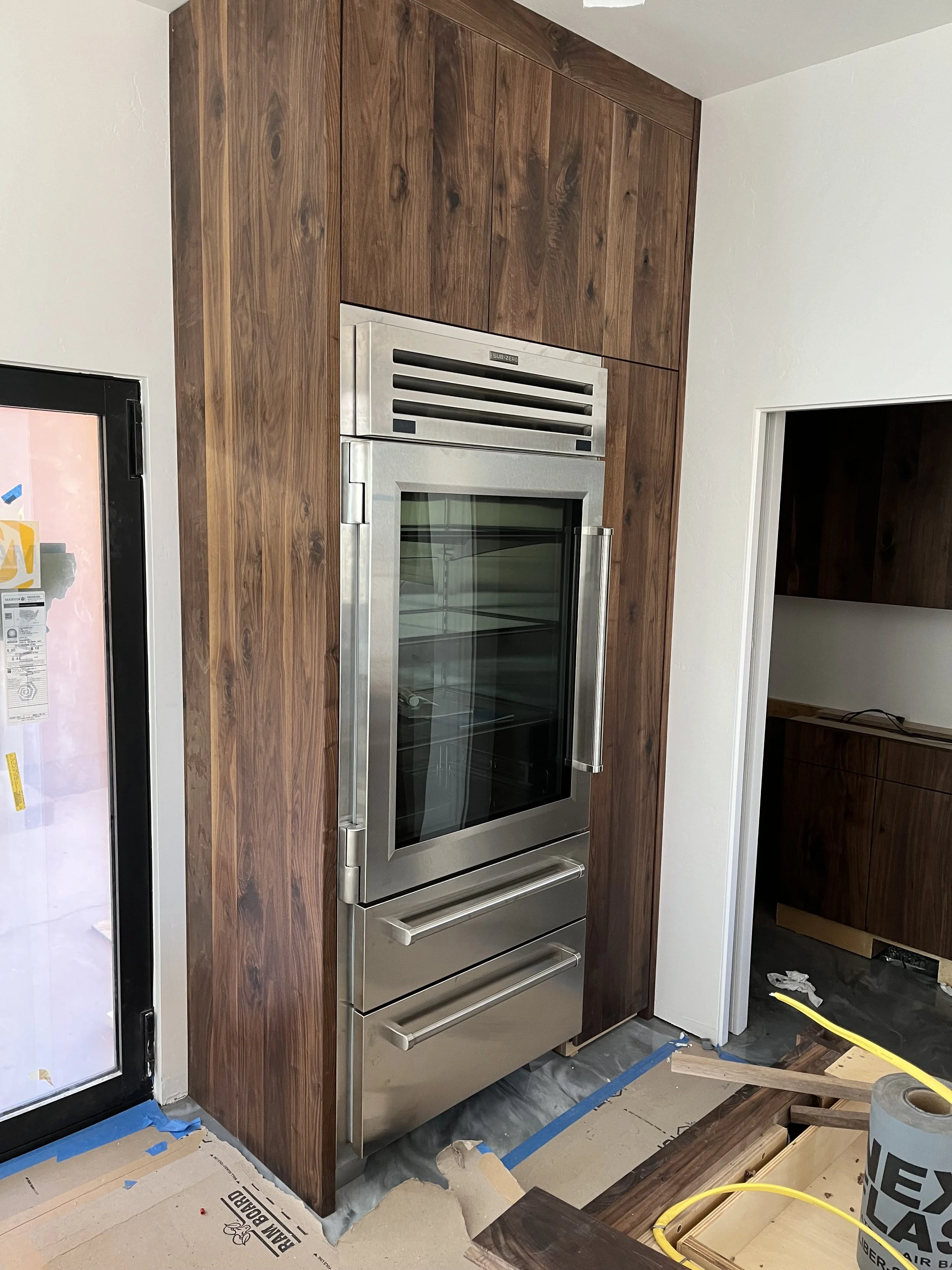 A commercial kitchen or food preparation area featuring a stainless steel oven or refrigerator built into a wooden cabinet wall, with partial view of a door on the left and some construction or renovation materials on the floor and nearby.