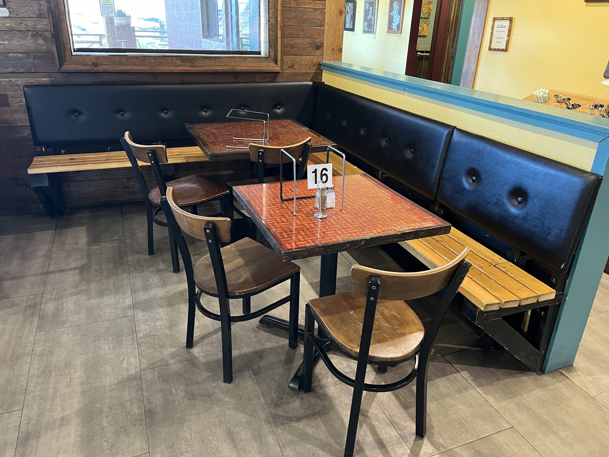 Corner of a restaurant with a dark blue upholstered bench, wooden tables and chairs, a window with a wooden frame, and wall art featuring colorful portraits.