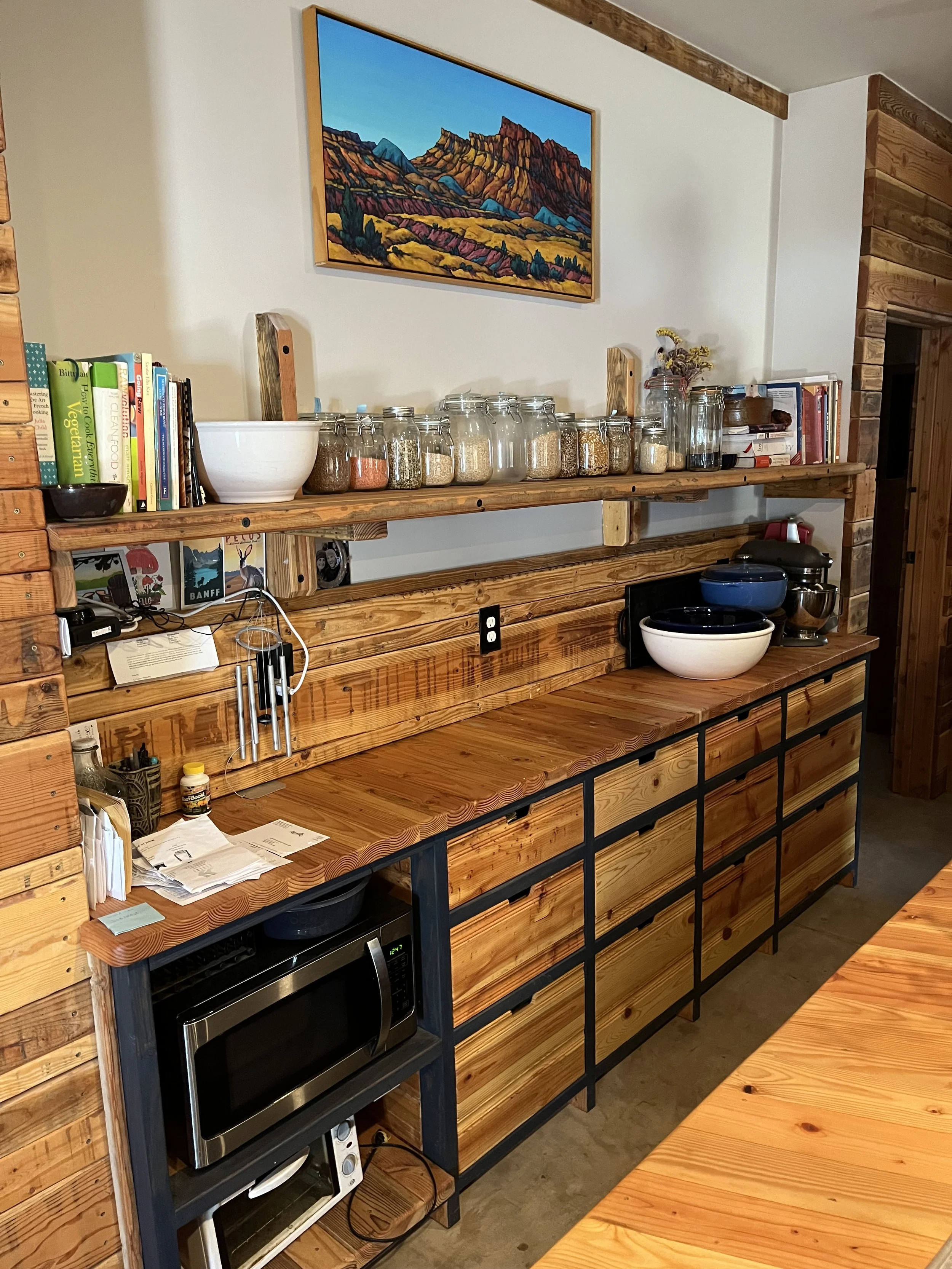 A rustic kitchen with wood accents and a long wooden shelf holding jars, books, and kitchen items. There is a countertop with a microwave oven and some bowls. The wall displays a colorful landscape painting of mountains.