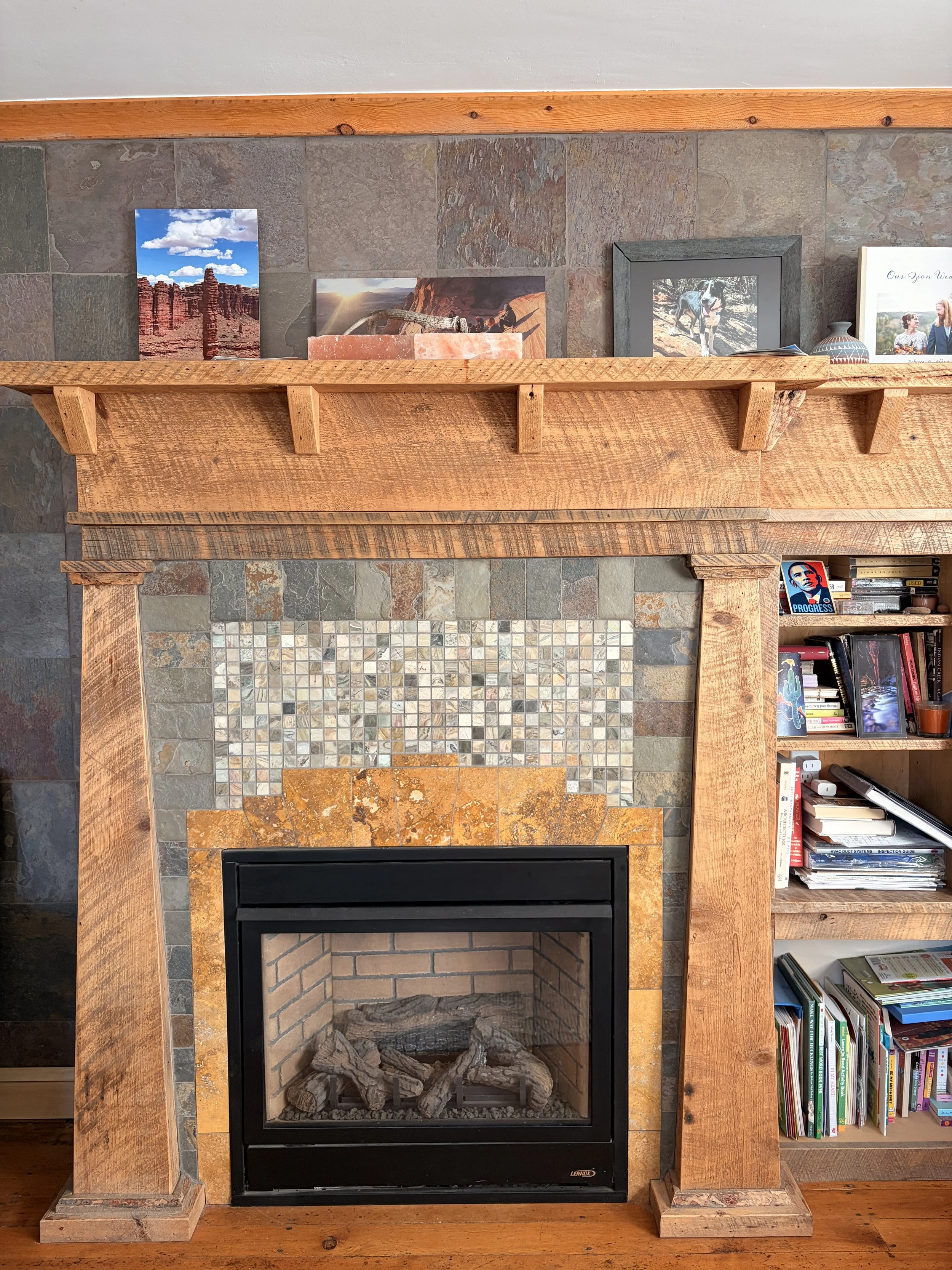 A fireplace with a wooden mantel and a tile surround, decorated with a framed photo of a dog, a landscape photo, a small vase, and a framed message. To the right, there is a bookshelf filled with books and some items.