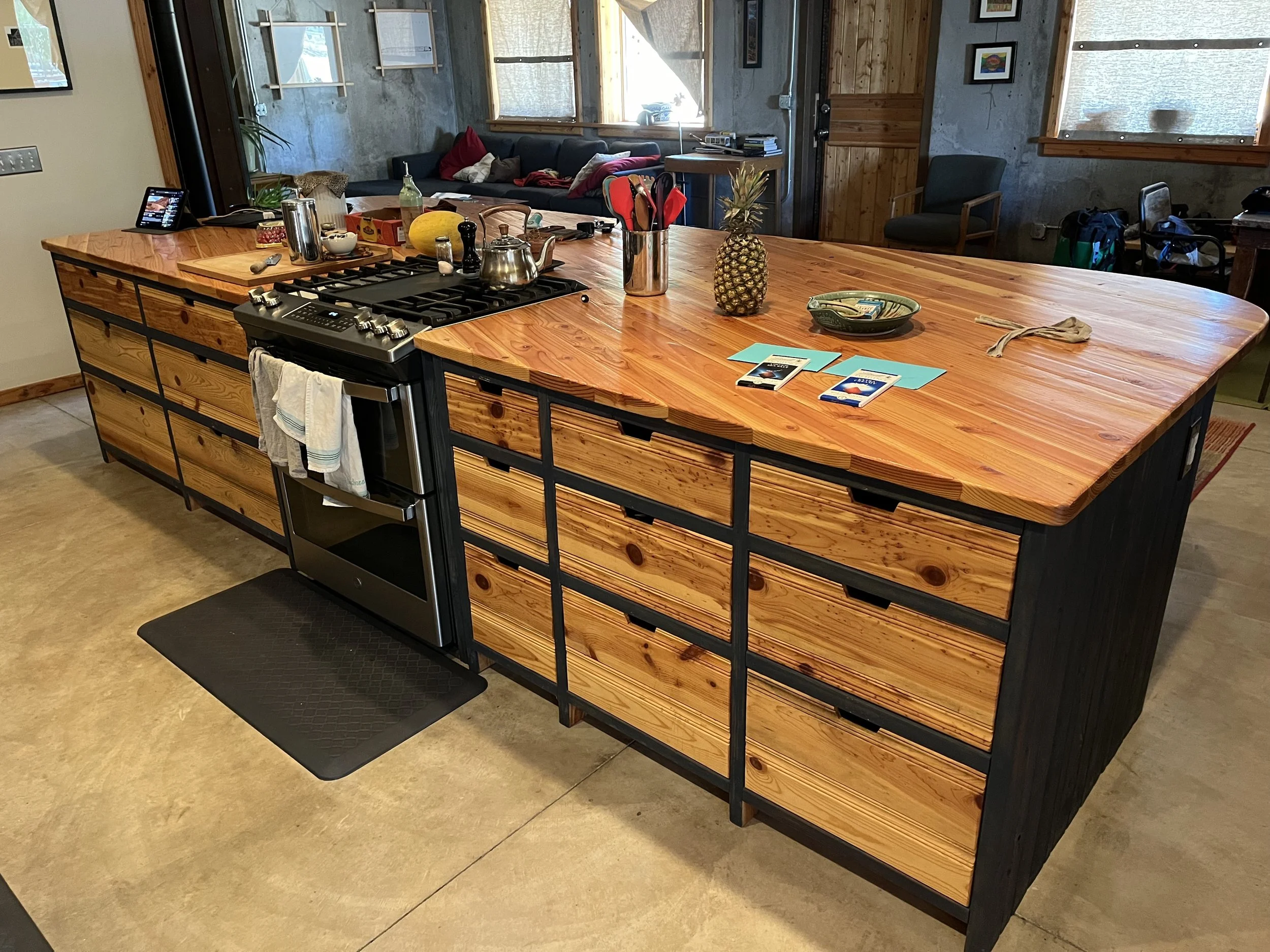 A large wooden kitchen island with a gas stove in the center, surrounded by various kitchen items including a pineapple, a bowl, and magazines. In the background, there is a living room with a black couch, several pillows, and windows letting in natural light.