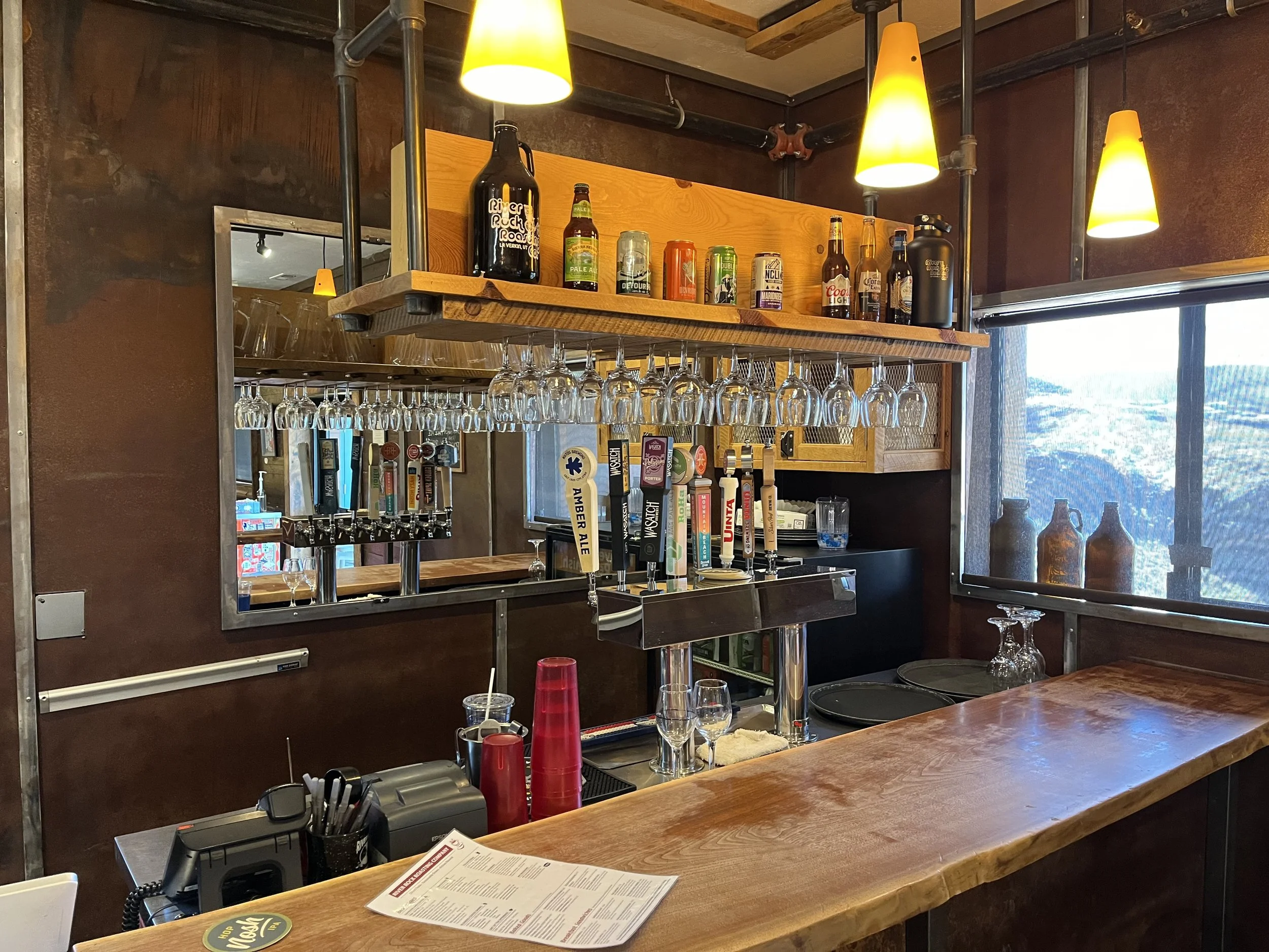Bar counter with beer taps, glasses hanging upside down, and various bottles and cans of beer on a shelf above. There are pendant lights hanging from the ceiling and a large window in the background.