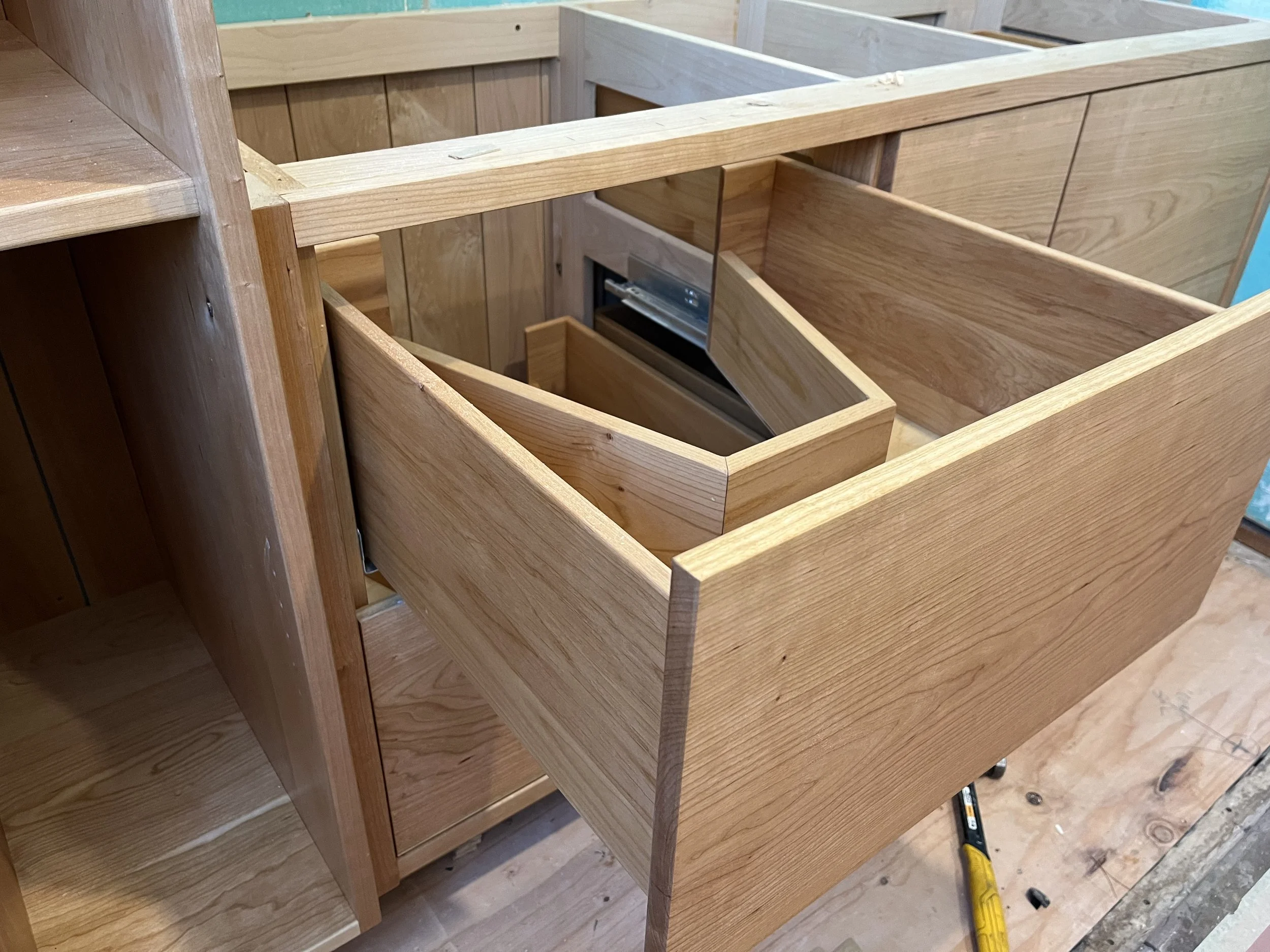 Wooden cabinets being assembled in a workshop, with a yellow screwdriver and a measuring tape on the floor.