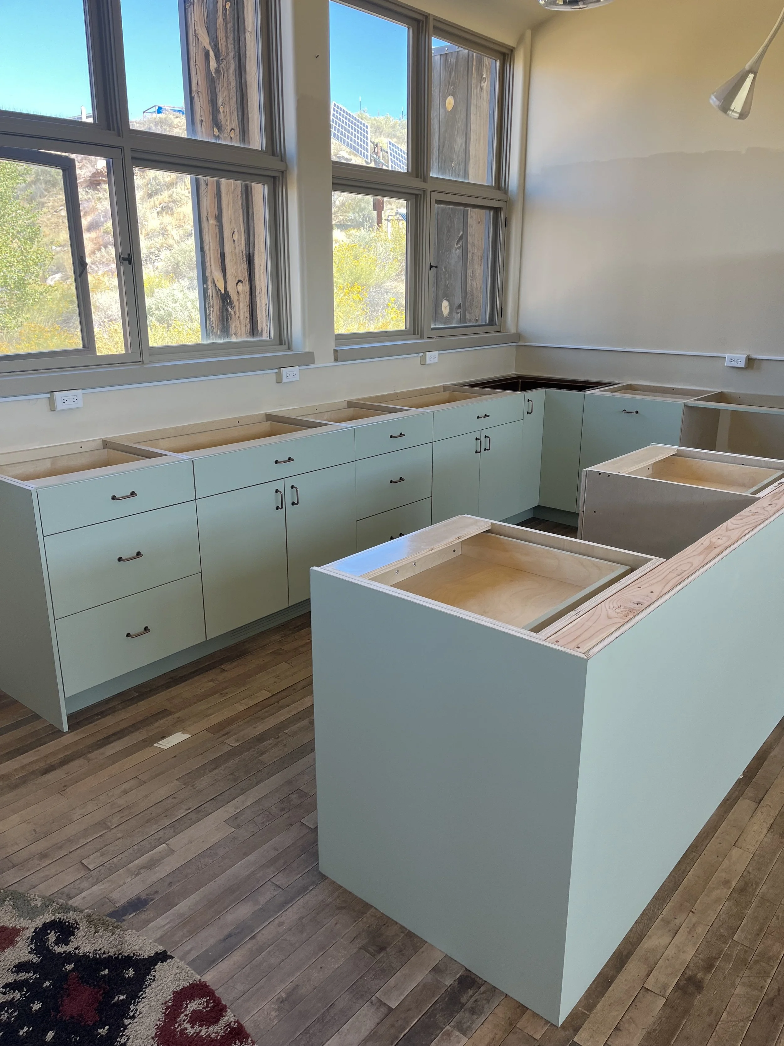 An unfinished kitchen with light green cabinets, some without countertops, next to large windows that let in natural light. There are electrical outlets along the walls, and the floor is wooden.