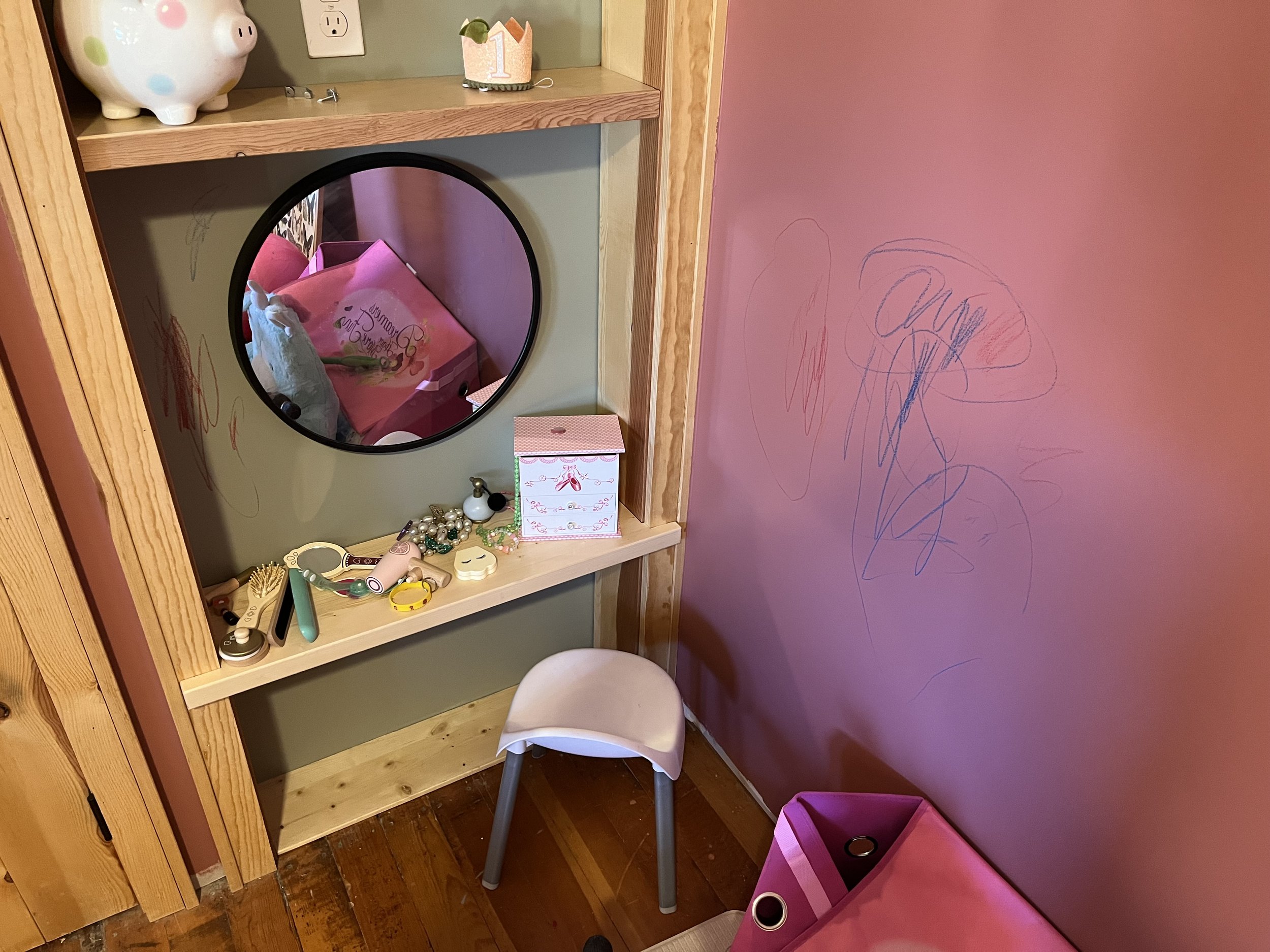 A children's vanity area with a mirror, pink walls, and various toys and jewelry scattered on a wooden shelf. There is a small chair in front of the vanity and a pink storage bin nearby.