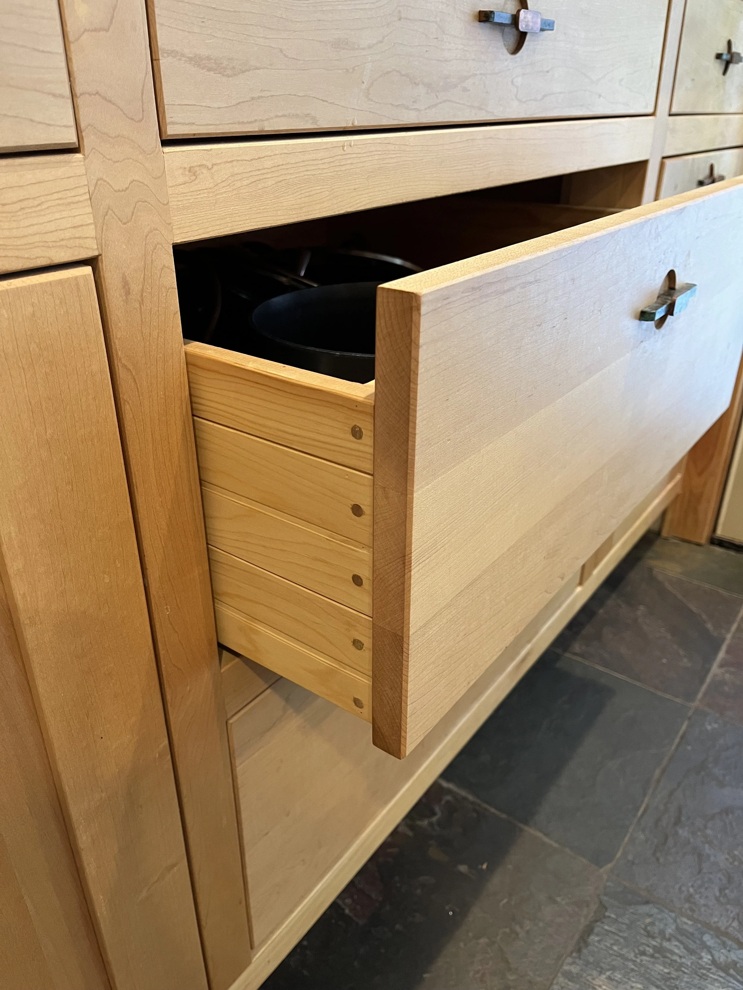 An open wooden drawer with a black pot inside, part of a wooden kitchen cabinet with multiple drawers, set on a dark tiled floor.