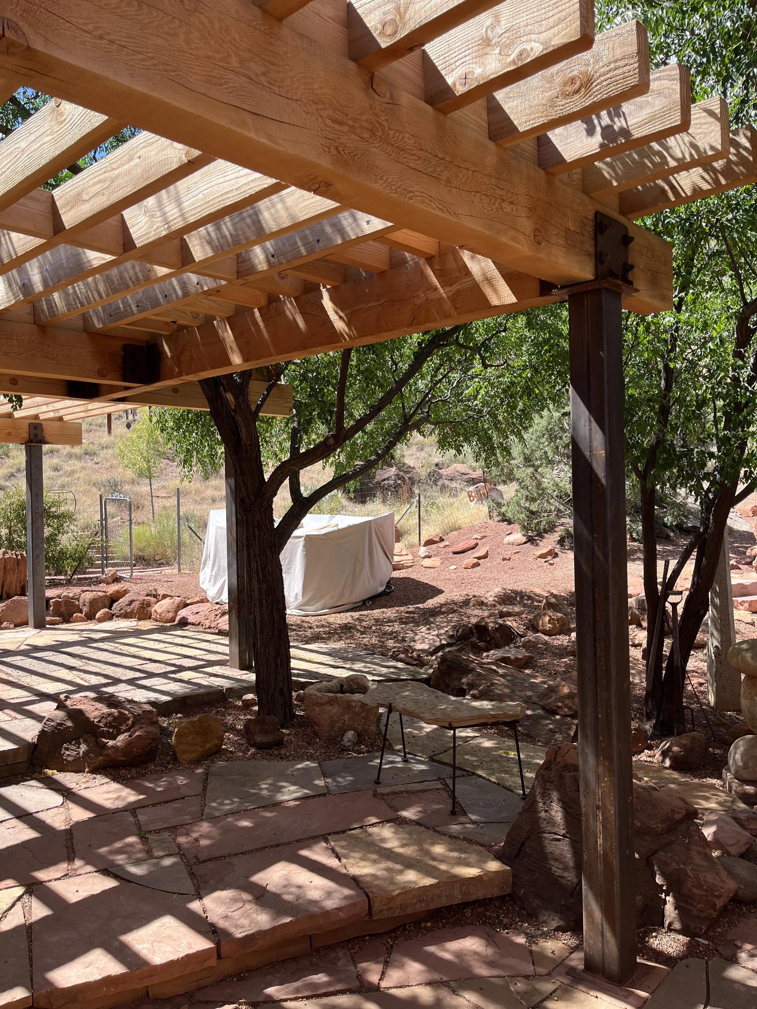 Wooden pergola with beams and slats, supporting overhead structure, shadow patterns cast on ground, surrounded by trees and rocks in a natural outdoor setting.