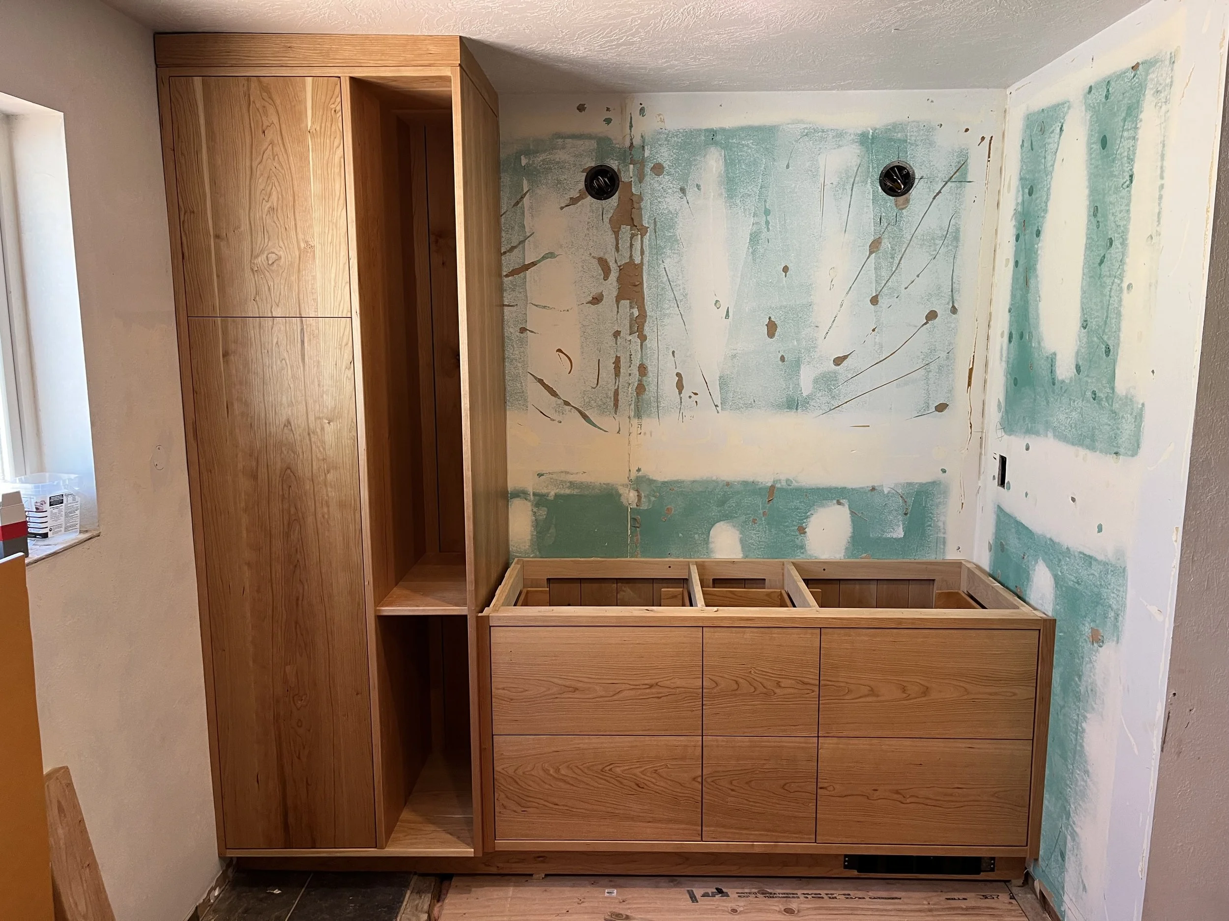 A wooden cabinet in the process of being installed in a room undergoing renovation. The wall behind has patches of drywall compound and paint splatters, with two round vents visible. Part of a window and some construction materials are also visible.