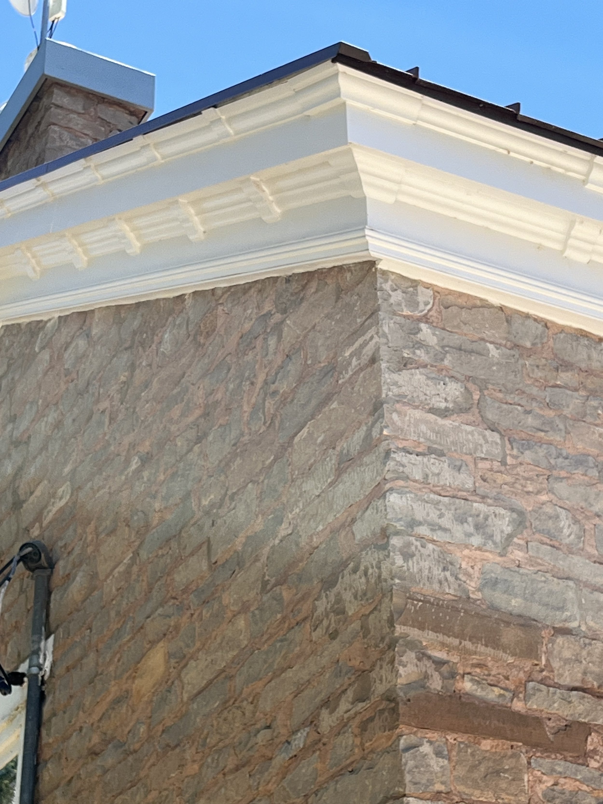 Corner of a house with stone walls, decorative white trim, and a roof with dark shingles, under a clear blue sky.