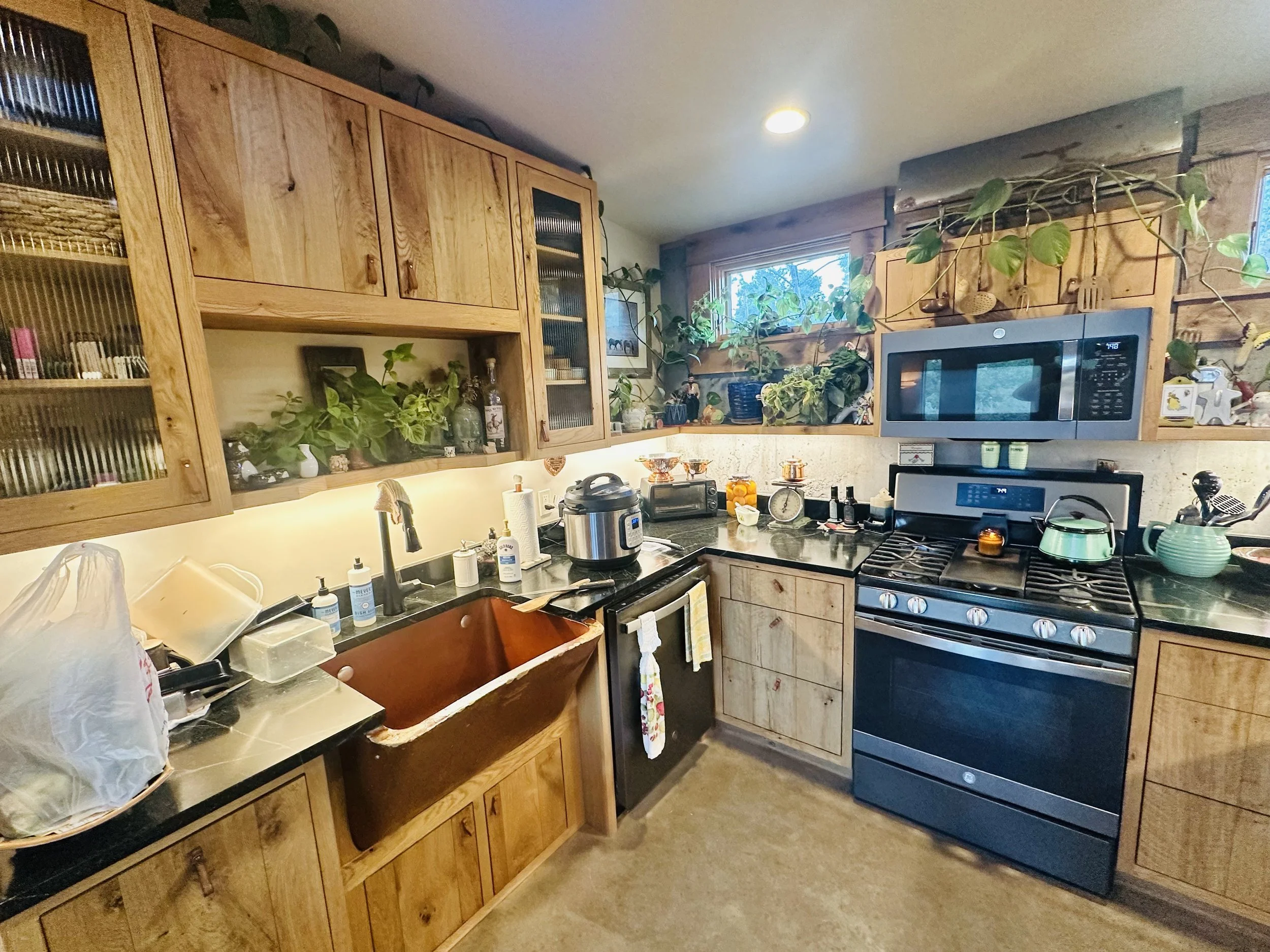 A cluttered kitchen with wooden cabinets, plants on the upper shelves, a microwave, stove, and various kitchen appliances and items on the countertops.