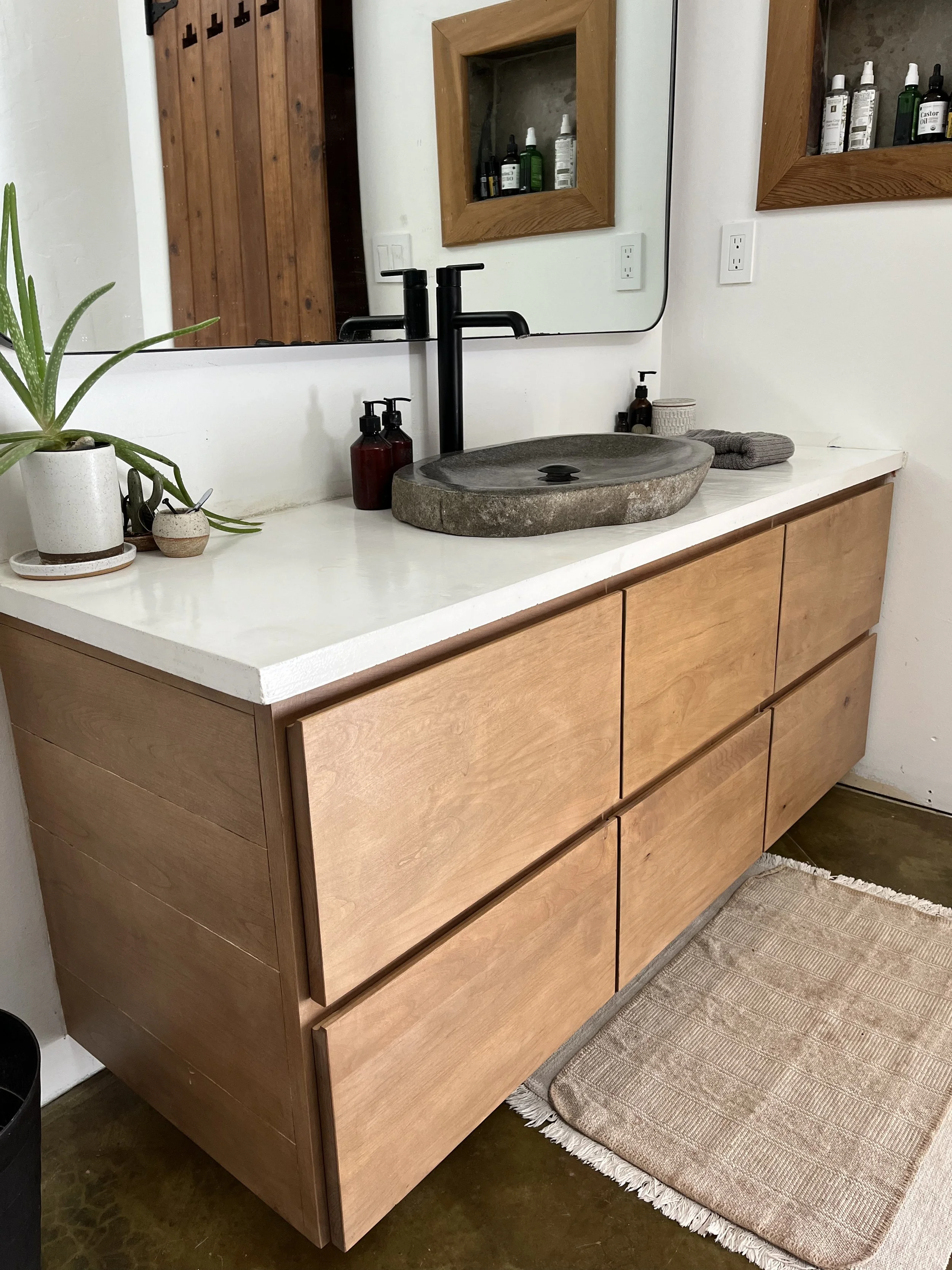 A modern bathroom vanity with a stone sink, black faucet, and wooden cabinet, decorated with potted plants, soap dispensers, and a towel, reflected in a mirror mounted above the sink.