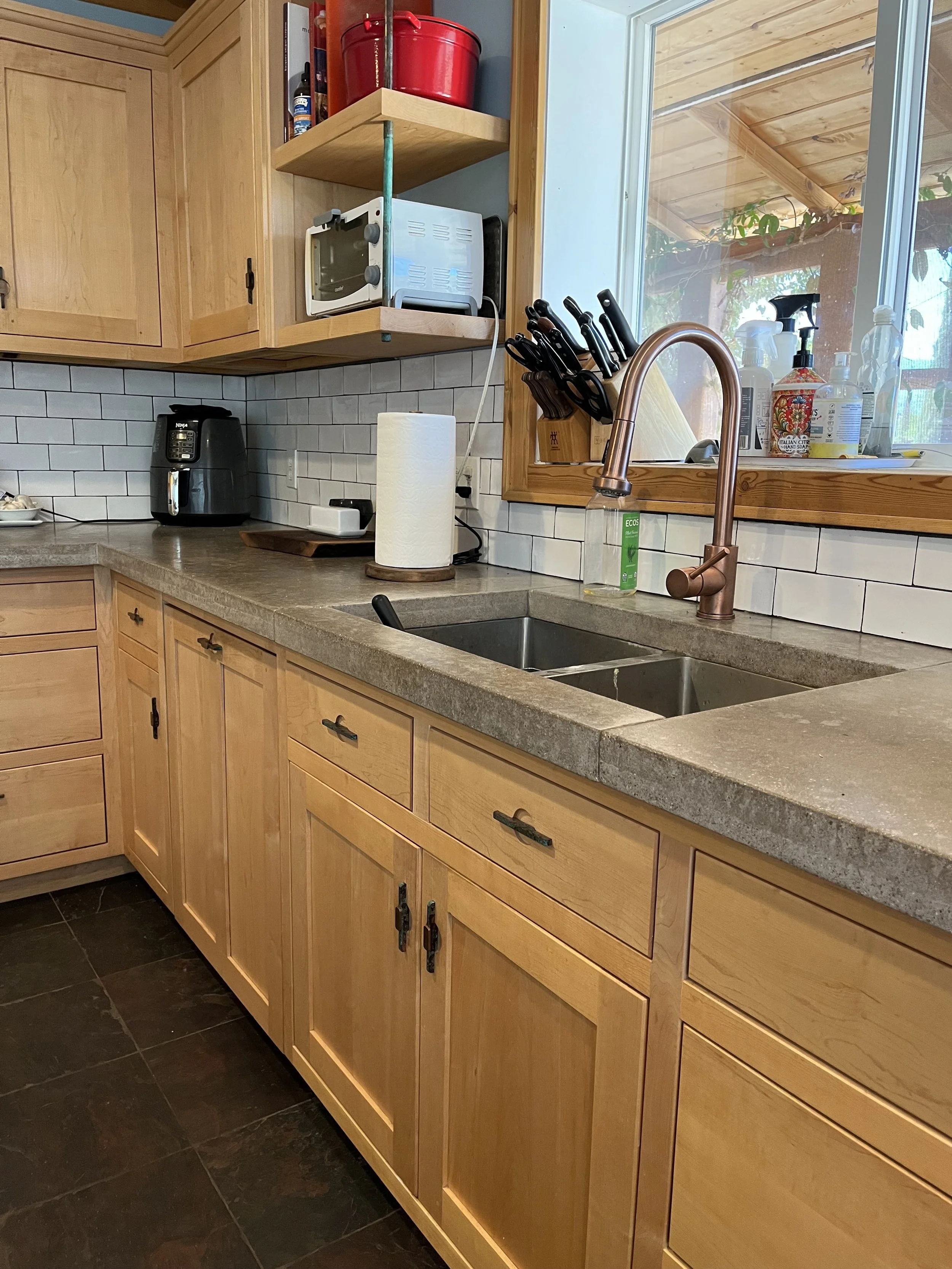 Kitchen with wooden cabinets, a countertop, a double sink, a rose gold faucet, and various kitchen appliances and supplies, including a paper towel roll, knives, soap bottles, and a microwave.
