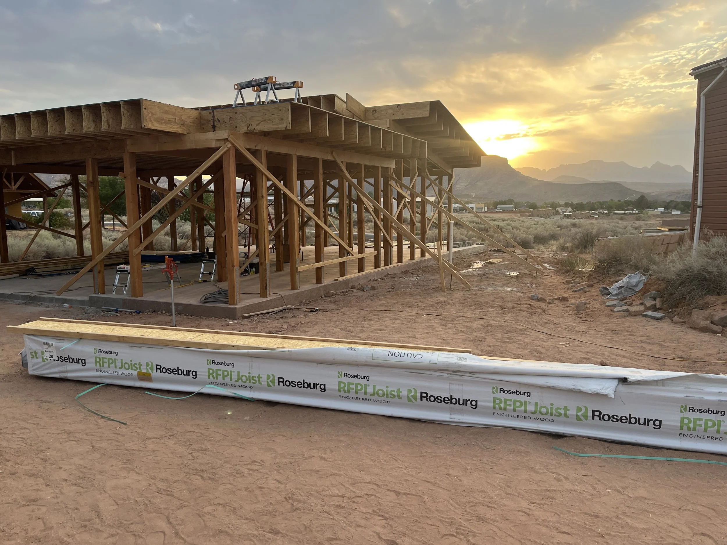 Construction site with wooden framework for a building, set against a sunset sky with mountains in the background. Building materials and tools are visible, along with a partially completed structure.