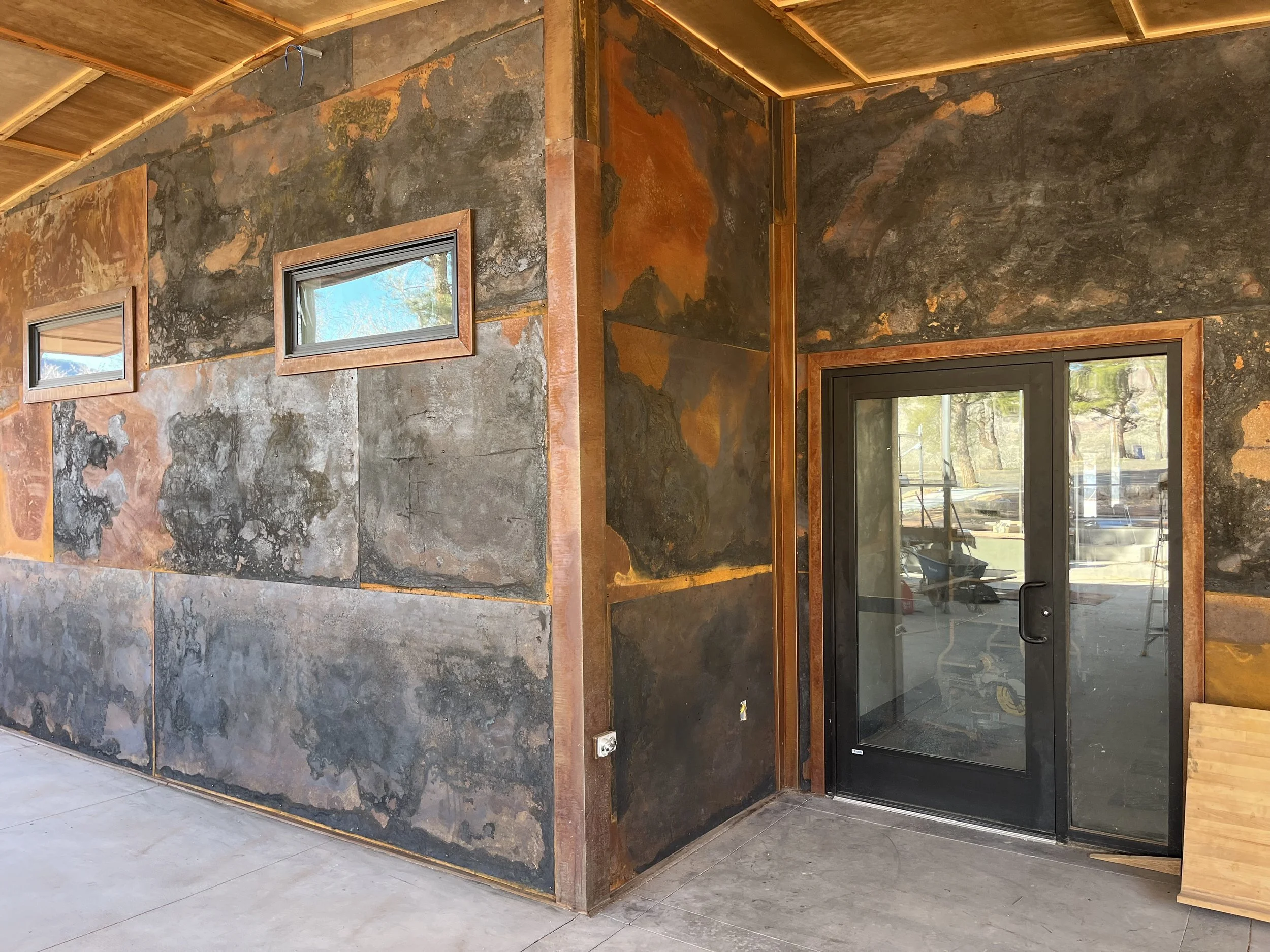 Interior of a modern building with natural stone walls, small horizontal windows, and wooden accents. The room features a glass door and an unfinished ceiling with exposed wiring.