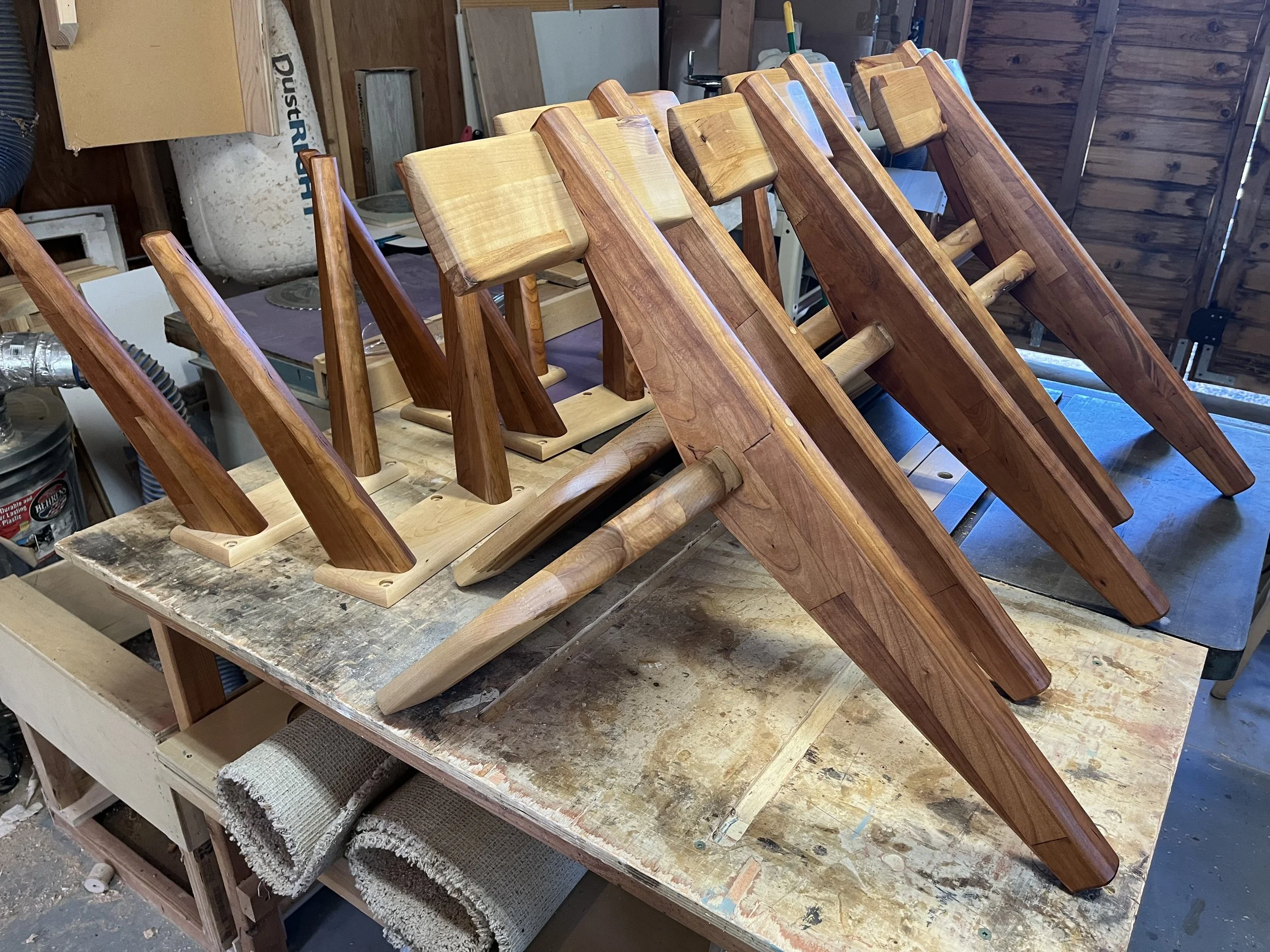 Several wooden chairs being assembled in a workshop, placed upside down on a workbench.