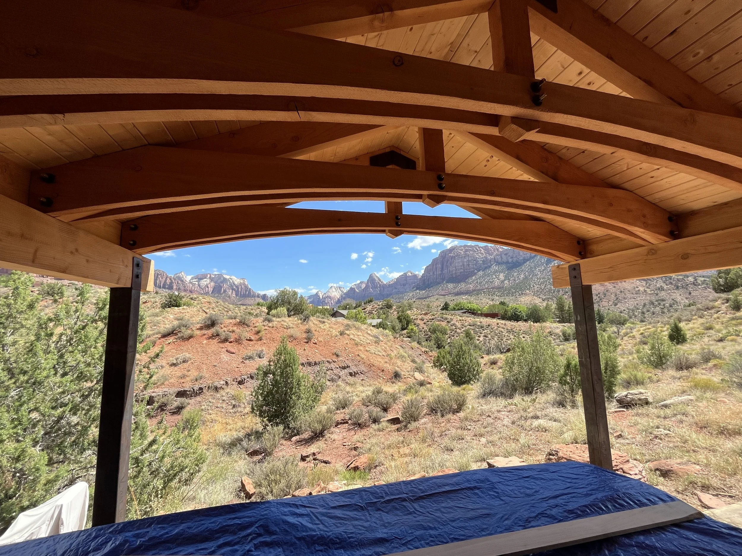 Landscape view from under a wooden structure, showing mountains, trees, and a partly cloudy sky in the background.