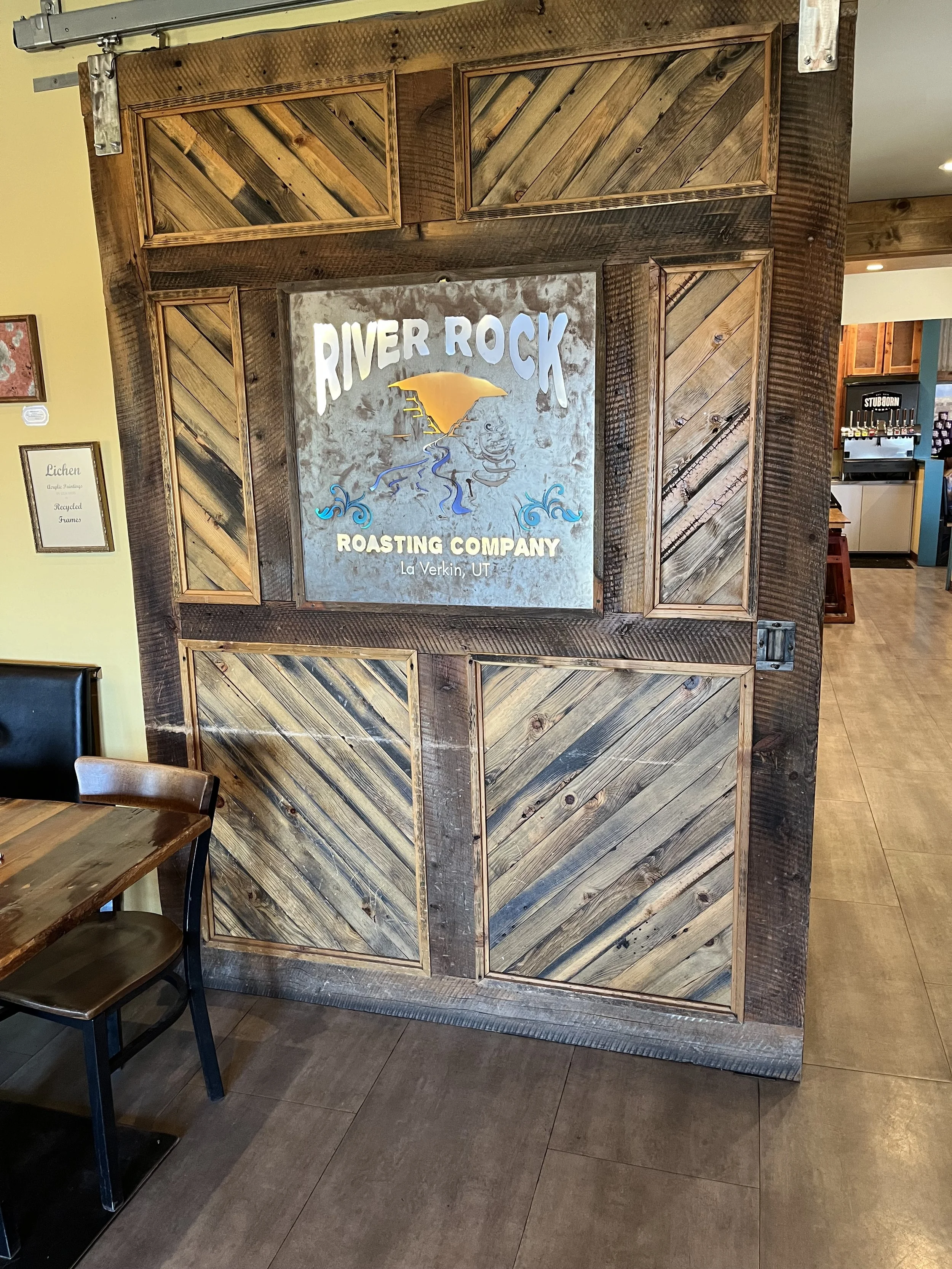 A wooden divider with a sign reading 'River Rock Roasting Company, La Verkin, UT' in a coffee shop, adjacent to a table and chairs, with a beverage station in the background.