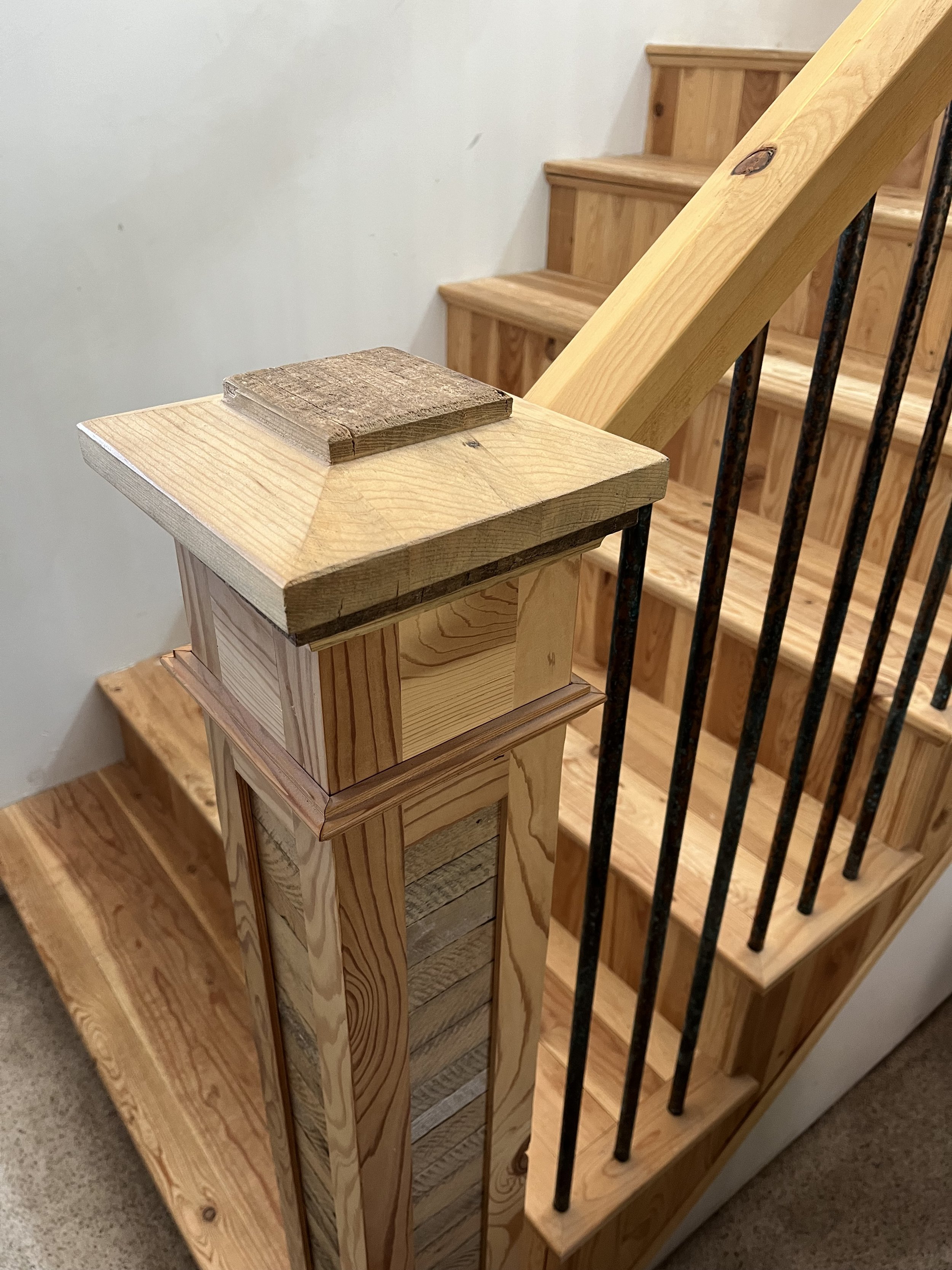 Close-up of a wooden staircase with new unfinished wood, featuring a wooden handrail, newel post, and black metal balusters.