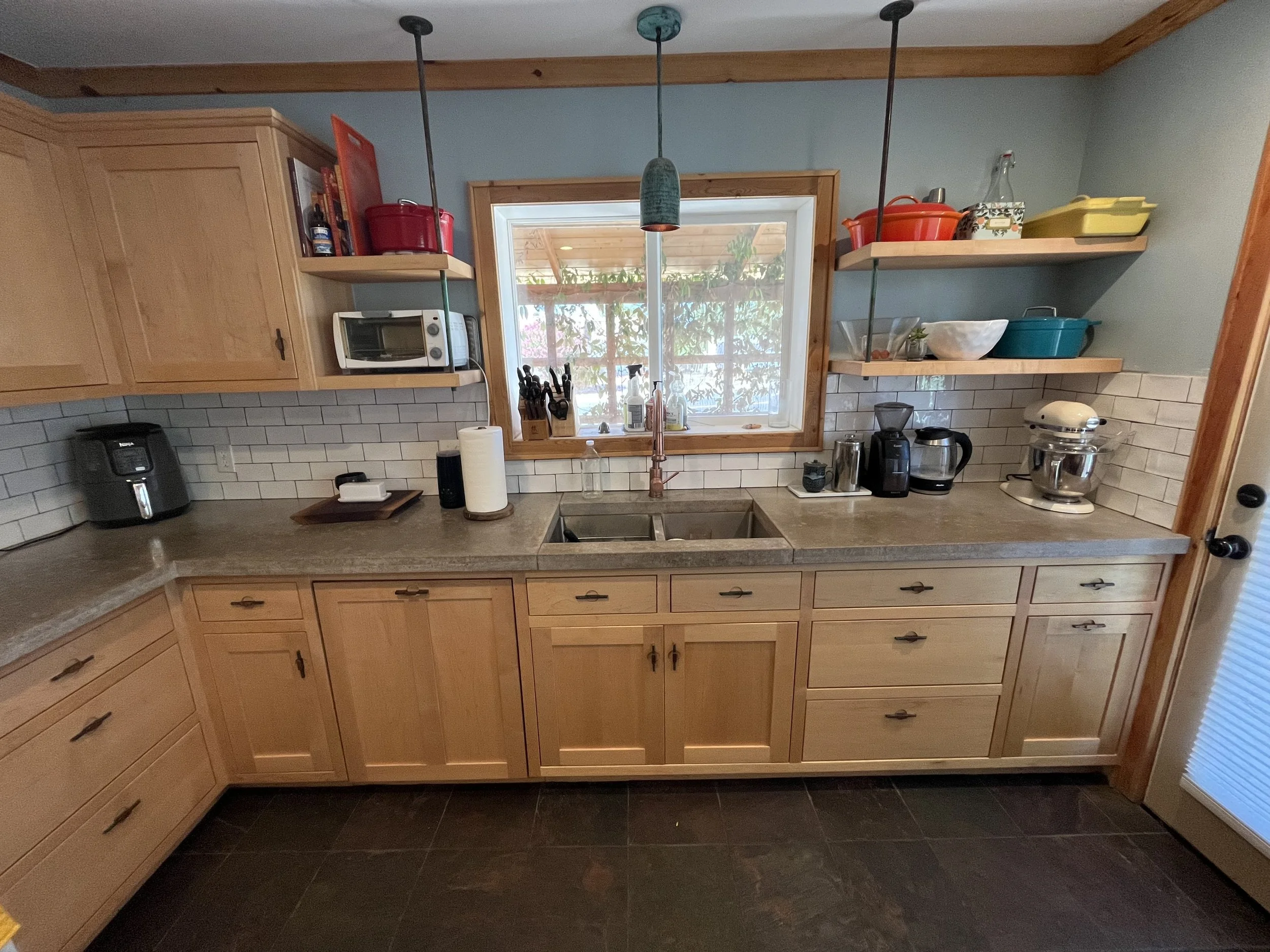 A kitchen with light wooden cabinets, a window above the sink, open shelves holding various kitchen items, and black floor tiles.