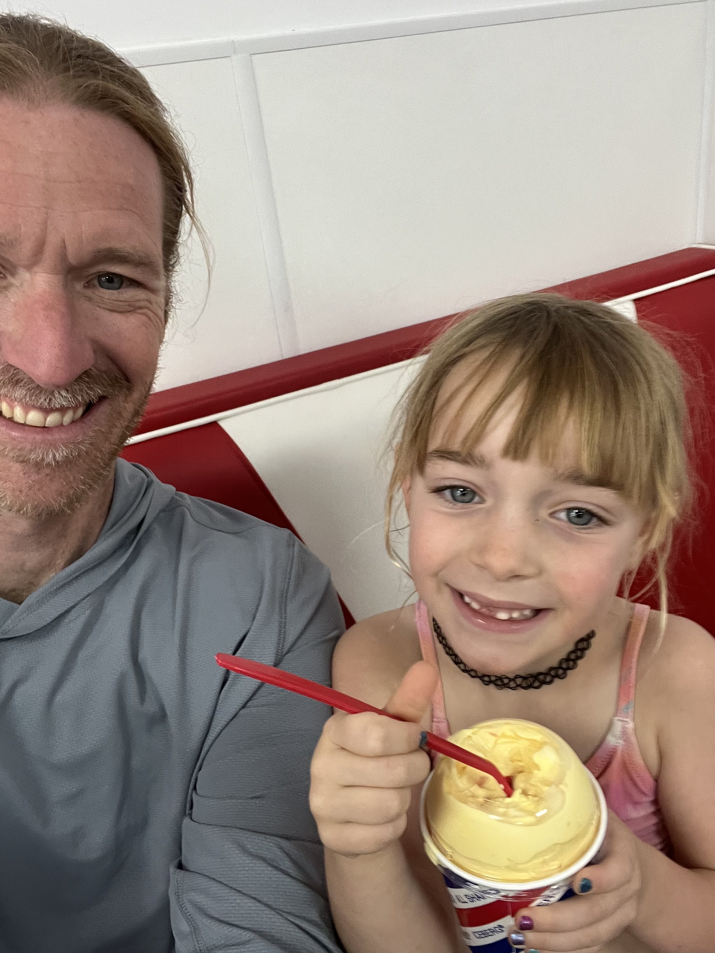 A man and a young girl smiling at the camera while holding a cup of yellow ice cream with a red spoon, in a room with white walls and a red cushioned seat.