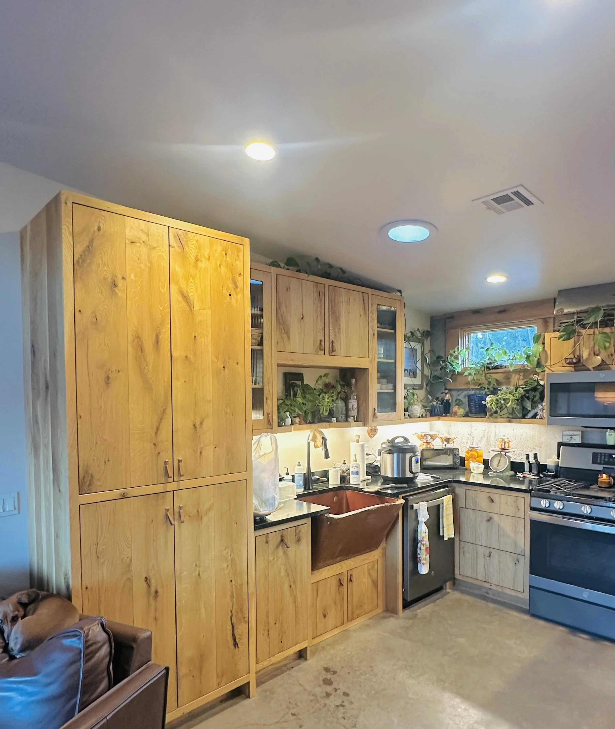 Kitchen with wooden cabinets, a microwave, a stove, plants on shelves, and a window with a view of trees outside.