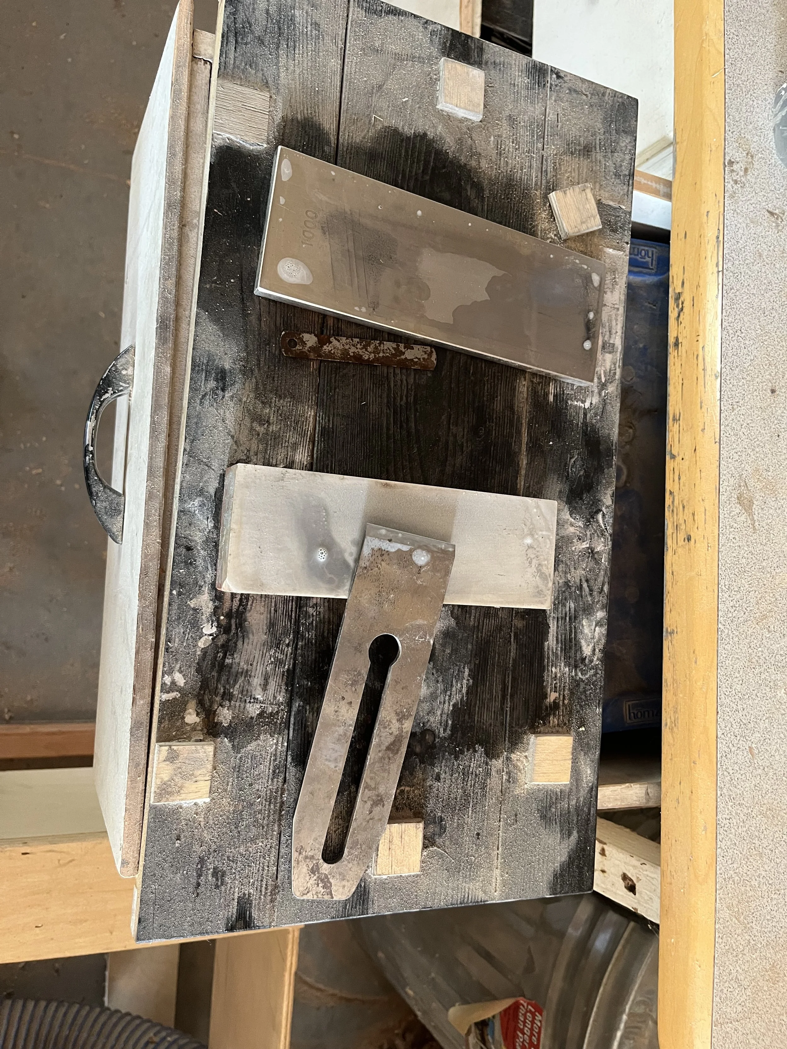 A workbench with a metal scraper, a rectangular metal object, and a stainless steel scale placed on a dirty, stained wooden surface in a workshop.
