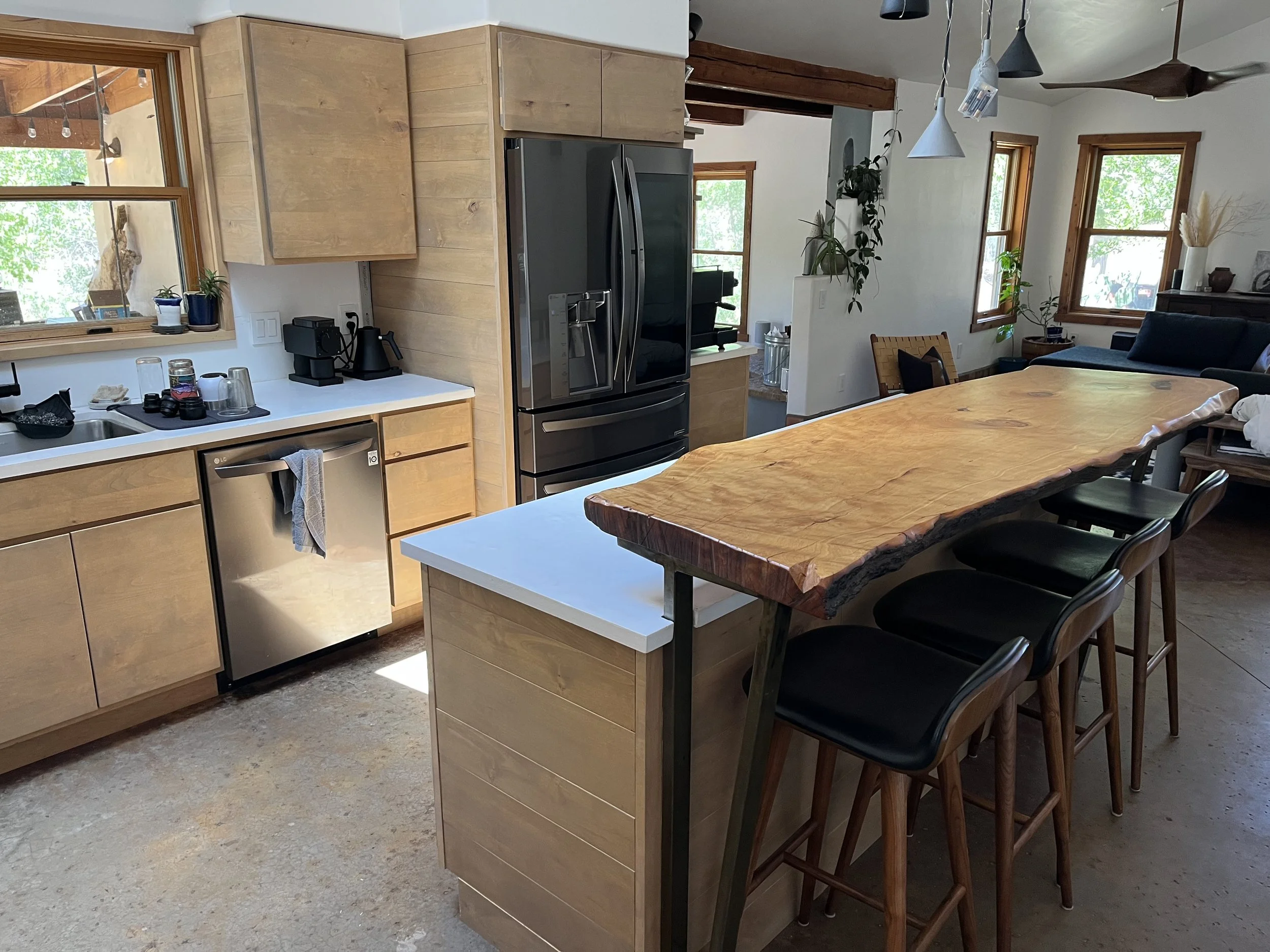 A modern kitchen and dining area with natural wood cabinets, a countertop with coffee maker and glasses, stainless steel refrigerator, wooden dining table with black chairs, and large windows letting in natural light.