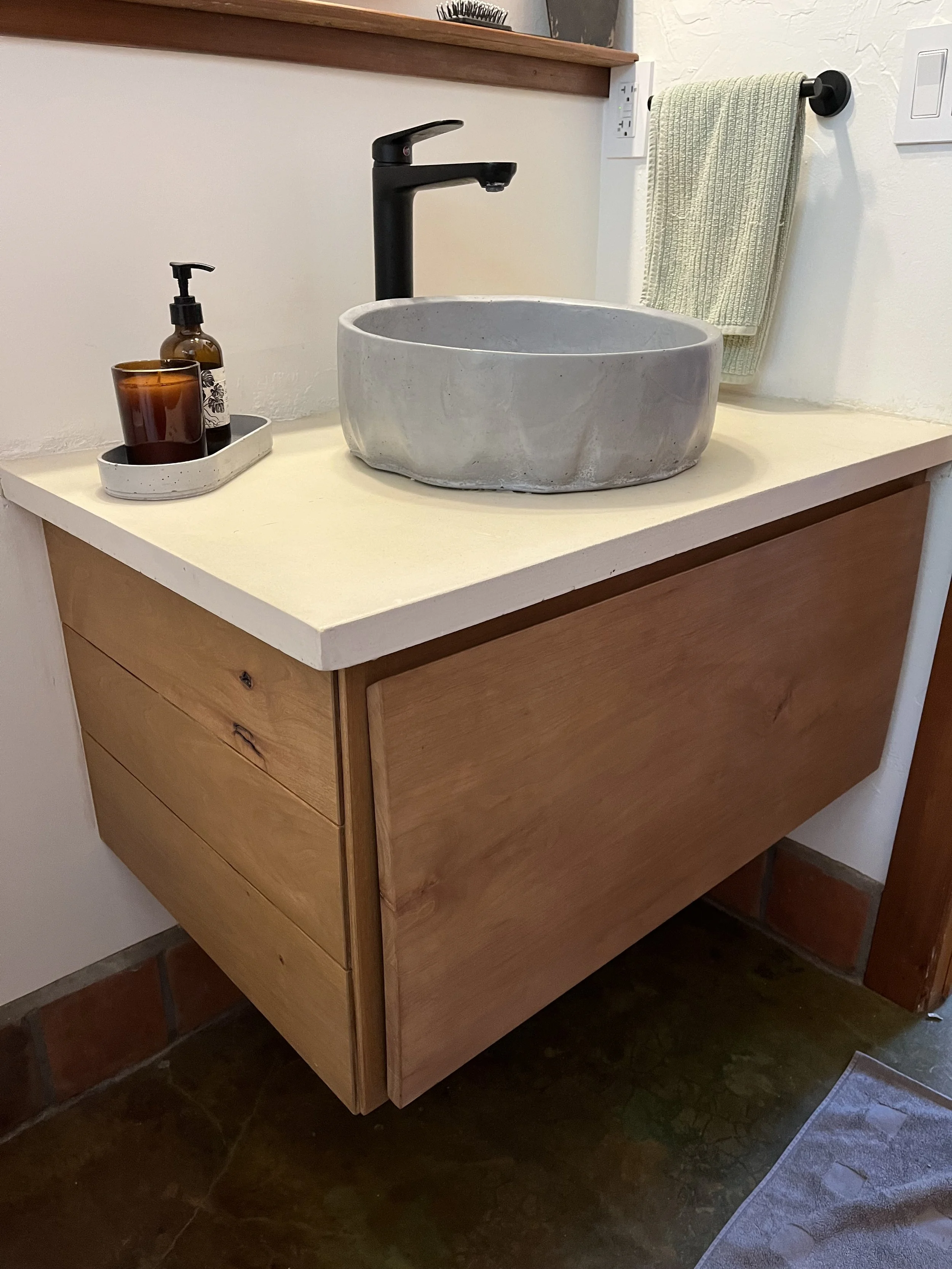 A bathroom vanity with a round stone sink and black faucet, a soap dispenser and candle, a light green towel hanging on a black towel bar, and part of a mirror with a wooden shelf above. The vanity has three drawers with visible wood grain, and the f