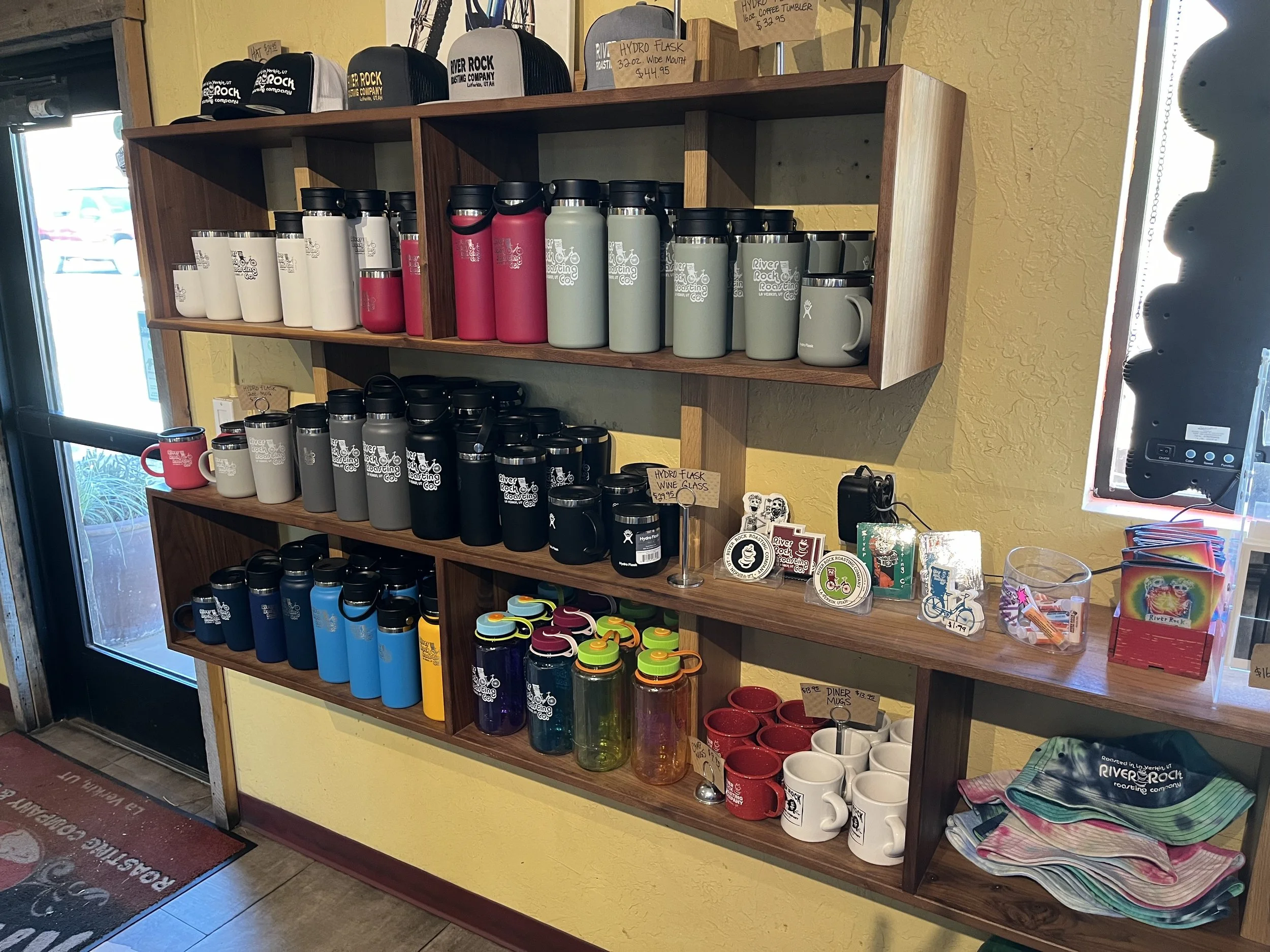 Display of various branded stainless steel water bottles and mugs on wooden shelves in a retail store. Some also have hats and other merchandise on top.