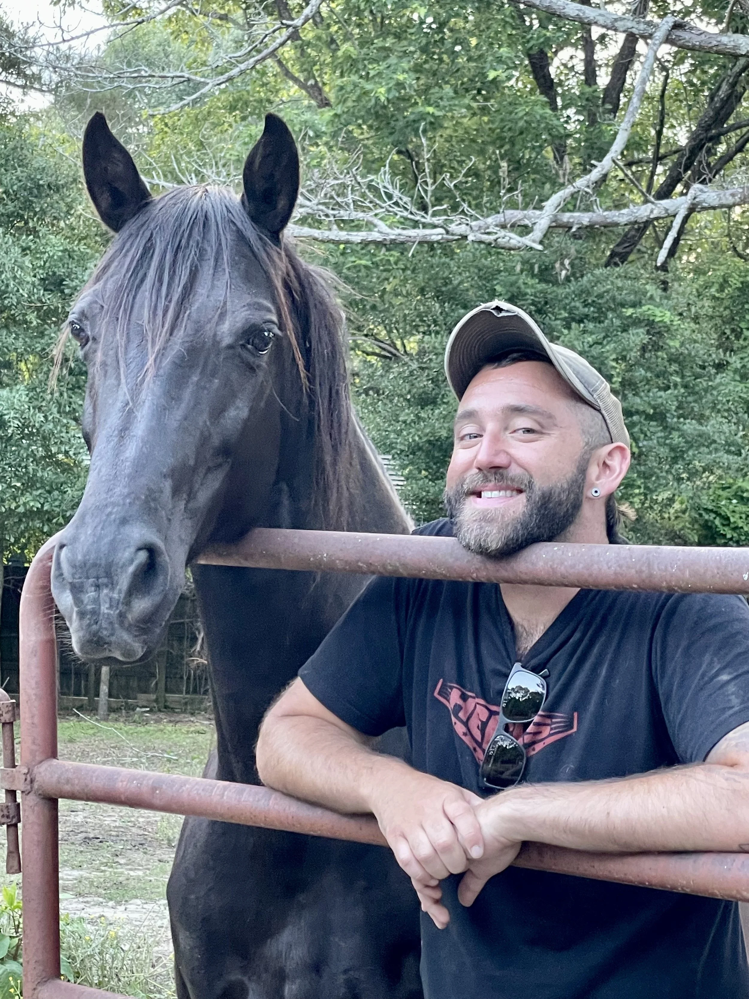 A smiling man with a beard and earrings, wearing a baseball cap and sunglasses hanging from his shirt, leans on a rust-colored metal fence. Behind him, a black horse with a long mane stands beside him, surrounded by green trees and foliage.