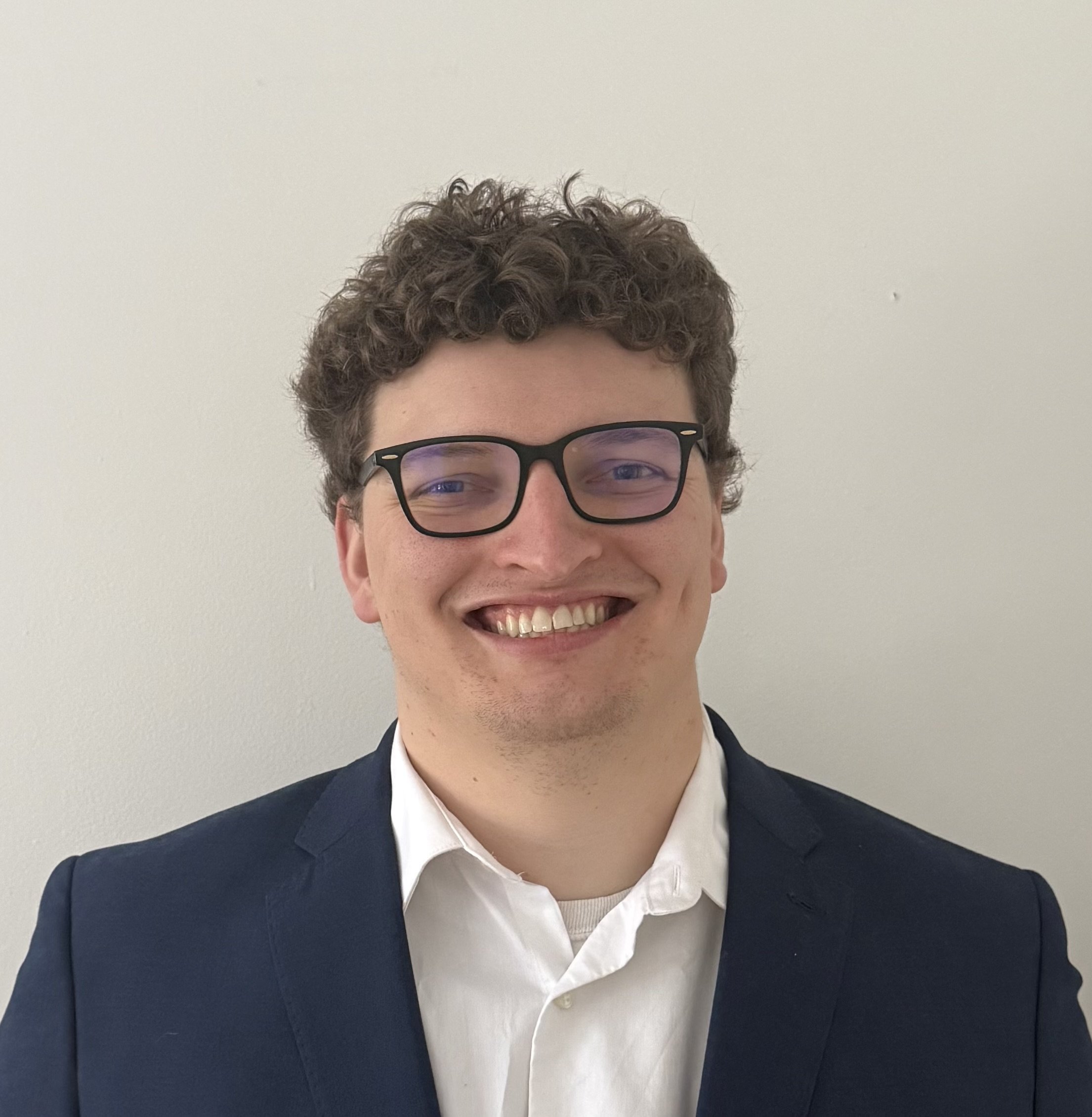 A smiling man with curly hair and glasses wearing a navy blazer and white shirt standing against a plain white wall.
