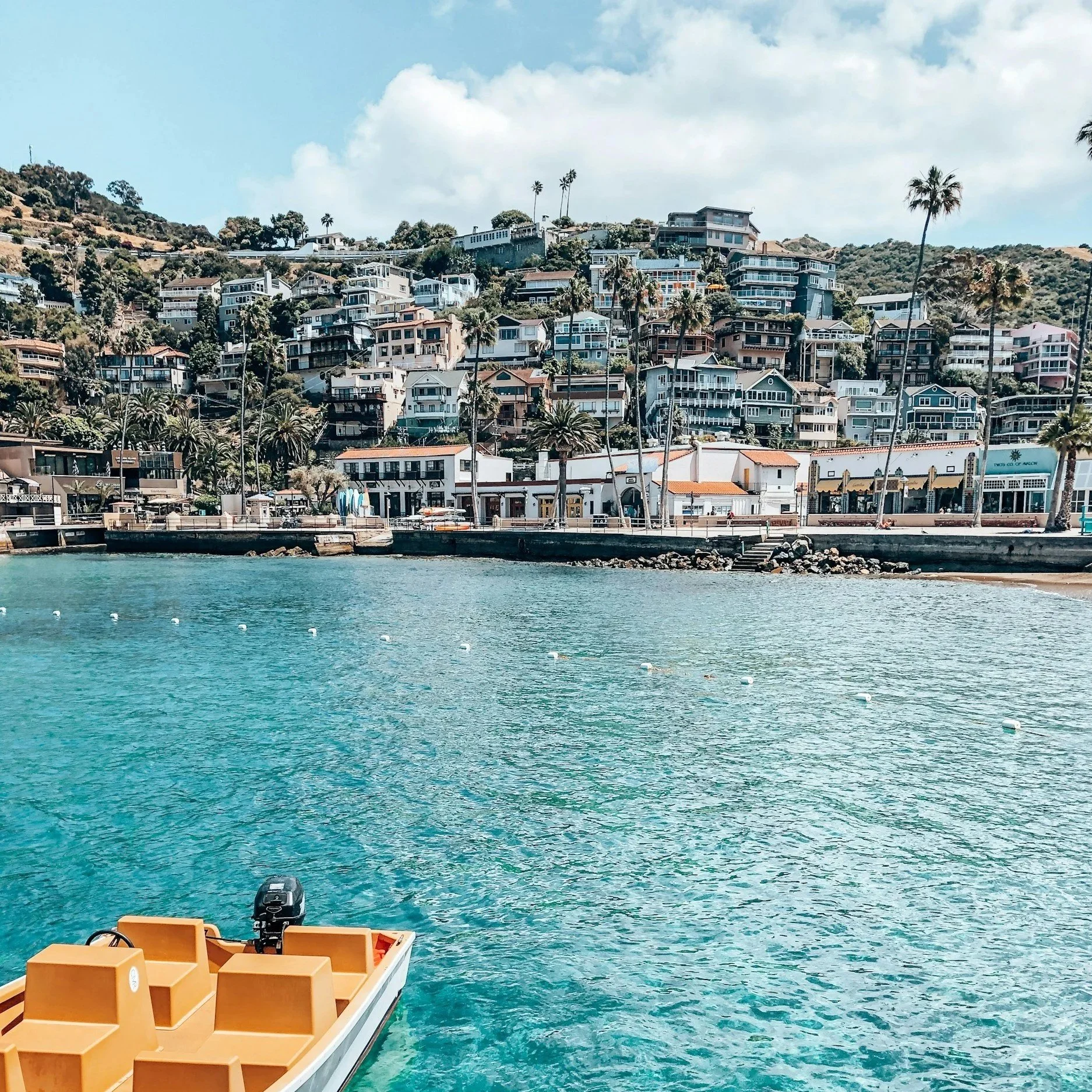 A coastal view with turquoise water, a small boat in the foreground, and an array of houses on a hillside in the background, along with palm trees and a partly cloudy sky.