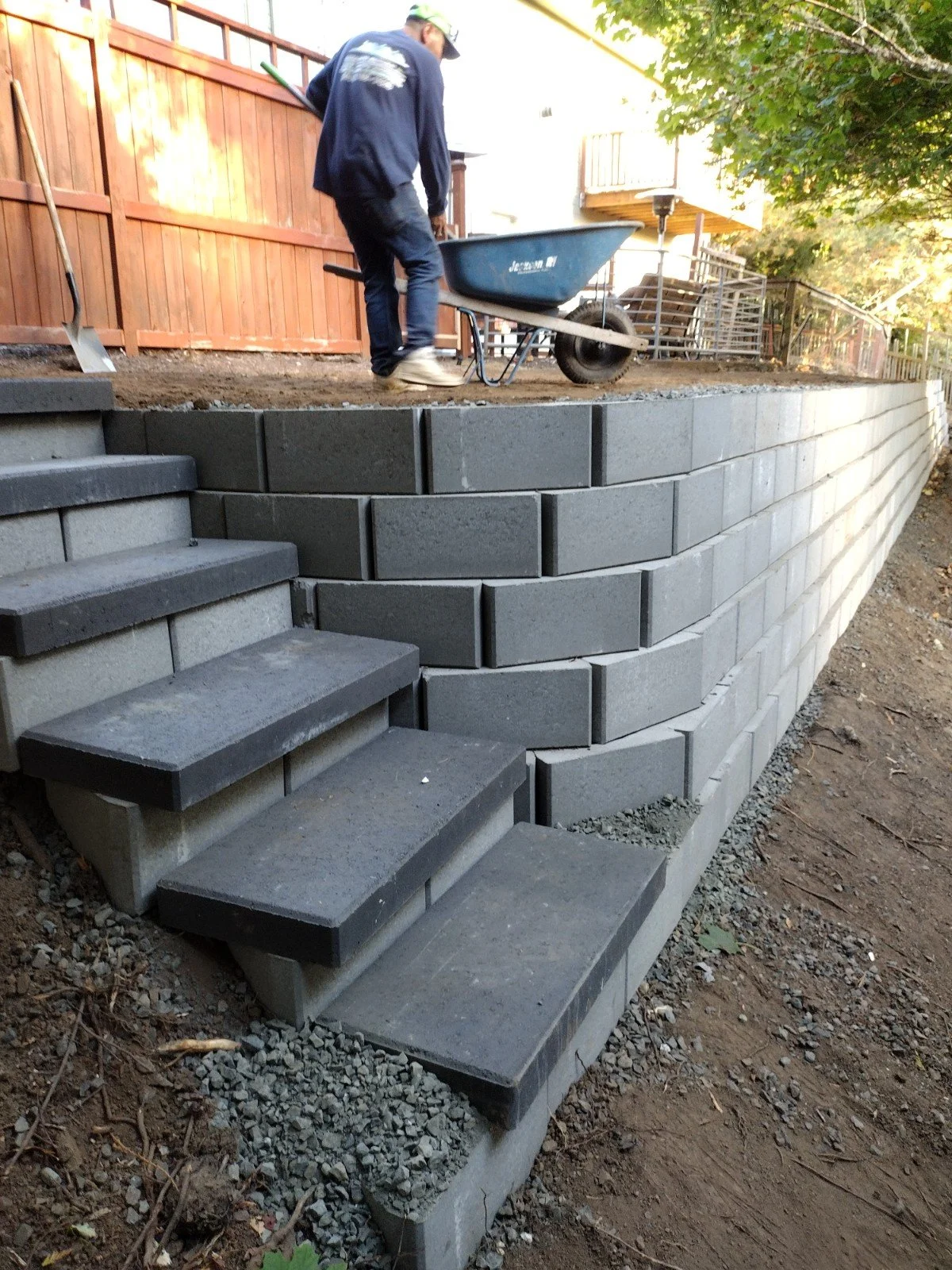 A construction worker is building a multi-layered concrete step and retaining wall system in a backyard. The worker is pushing a wheelbarrow filled with gravel or soil. The new wall features curved sections, and there are older concrete steps leading to the upper yard, which is enclosed by a wooden fence. Trees with green leaves are visible in the background.