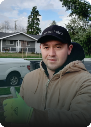 A man wearing a black cap, beige hoodie, and green gloves taking a selfie outdoors with a house and trees in the background.