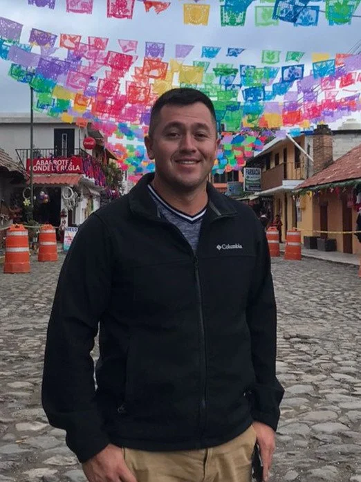 A man standing on a cobblestone street under colorful paper decorations hanging overhead, with buildings and orange barriers in the background.