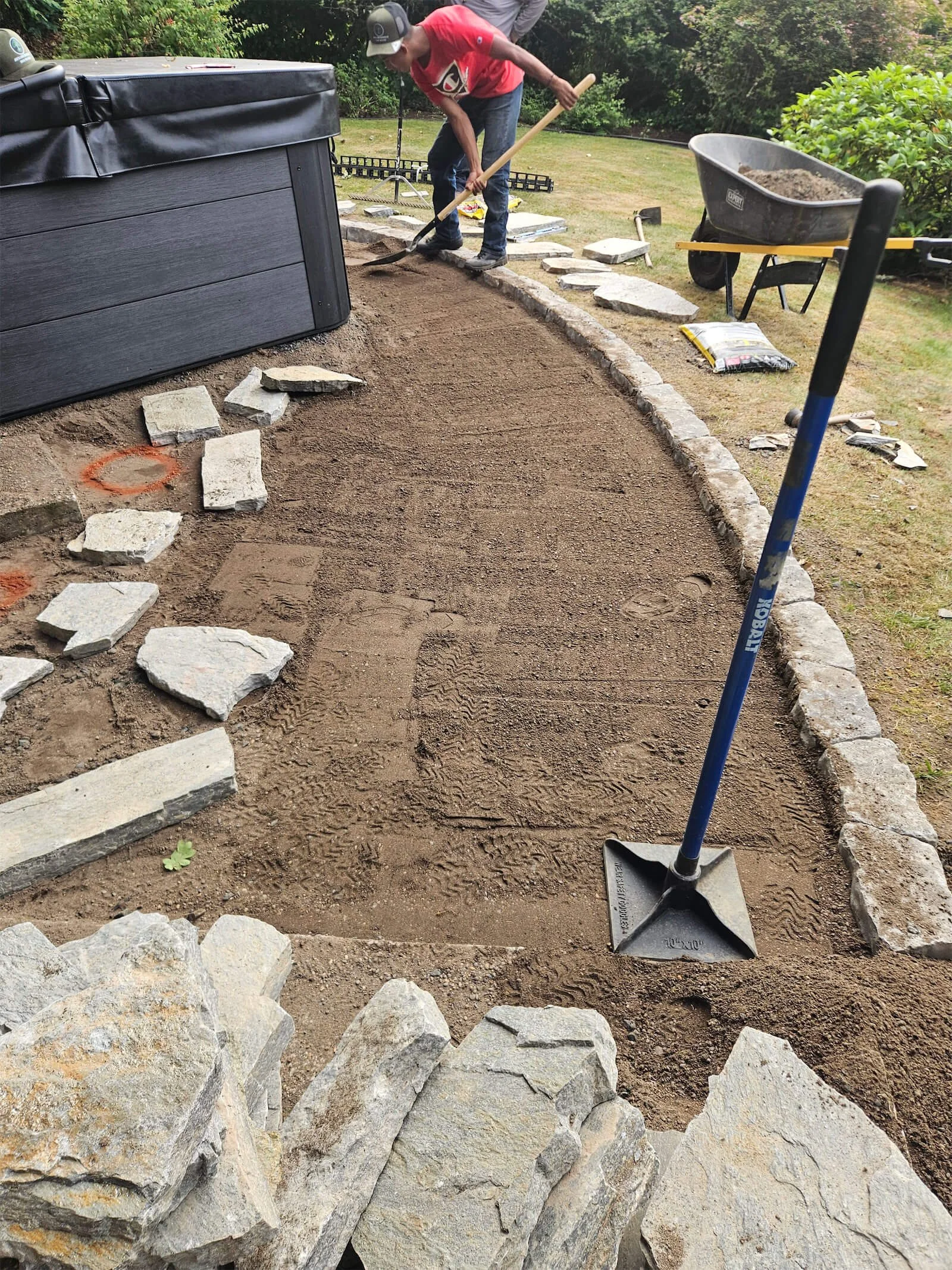 Person working on a landscaping project, installing a curved stone border with a shovel, wheelbarrow, and paving stones around a yard.