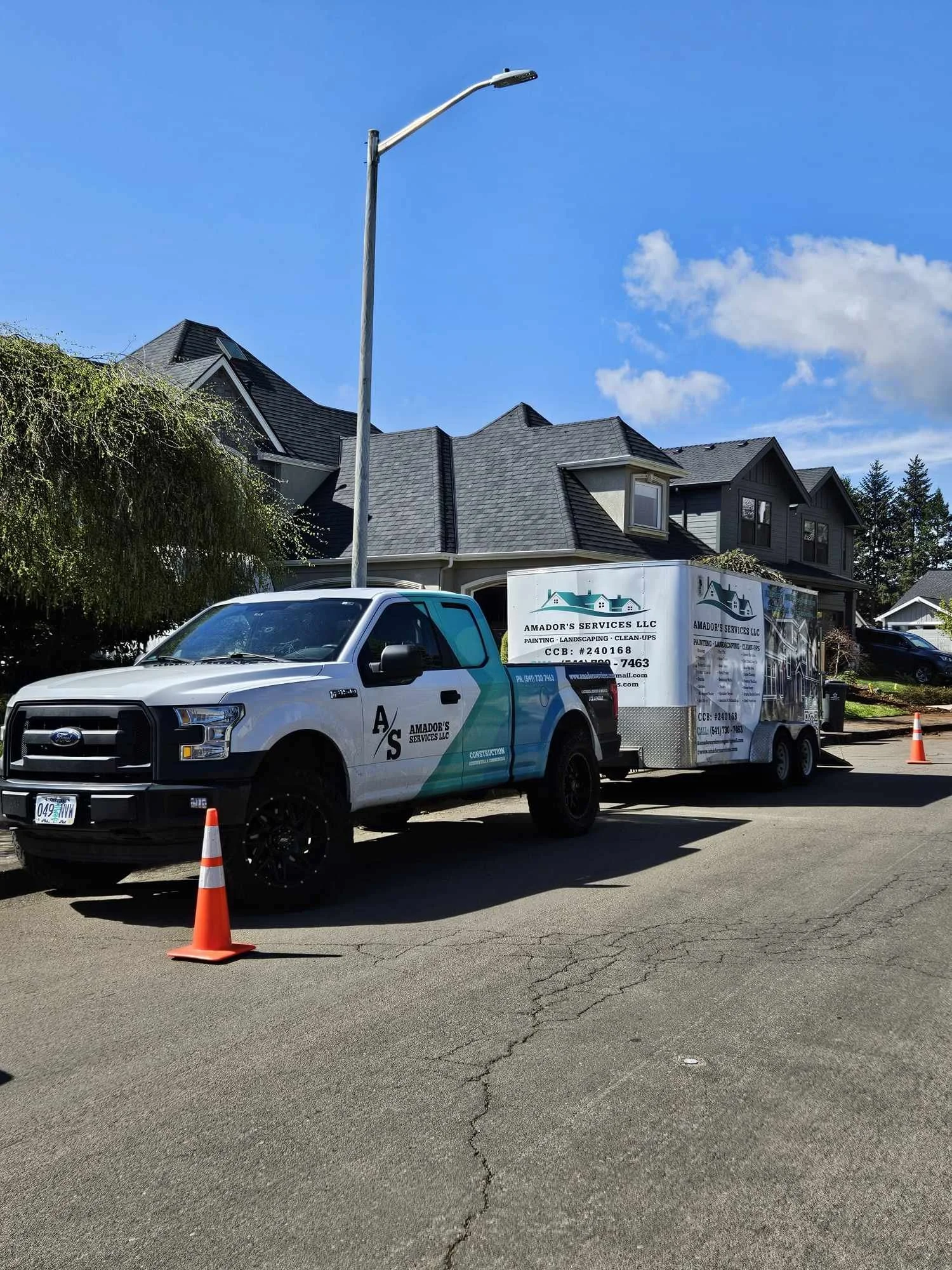 A white and teal Ford pickup truck with a trailer attached, parked on a street with orange traffic cones nearby. The trailer displays the name 'Amador's Services LLC' and services listed. The scene is in a residential neighborhood with houses and a clear blue sky.