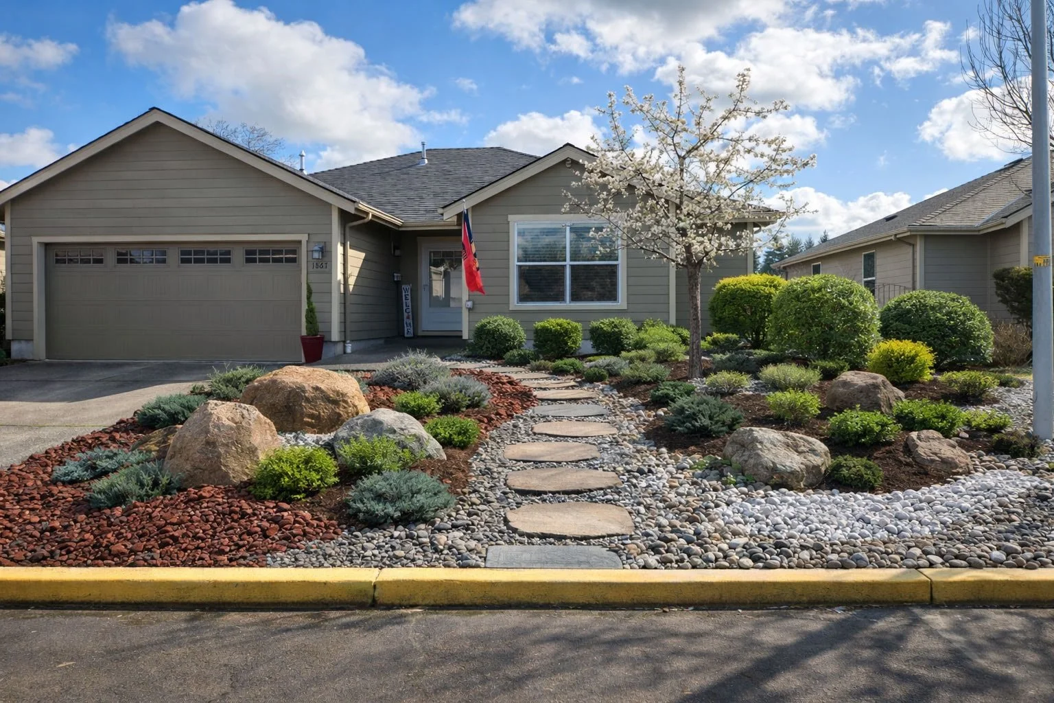 Front yard landscaping of a house with a stone path, rocks, and various green shrubs, a small flowering tree, and an American flag near the entrance.