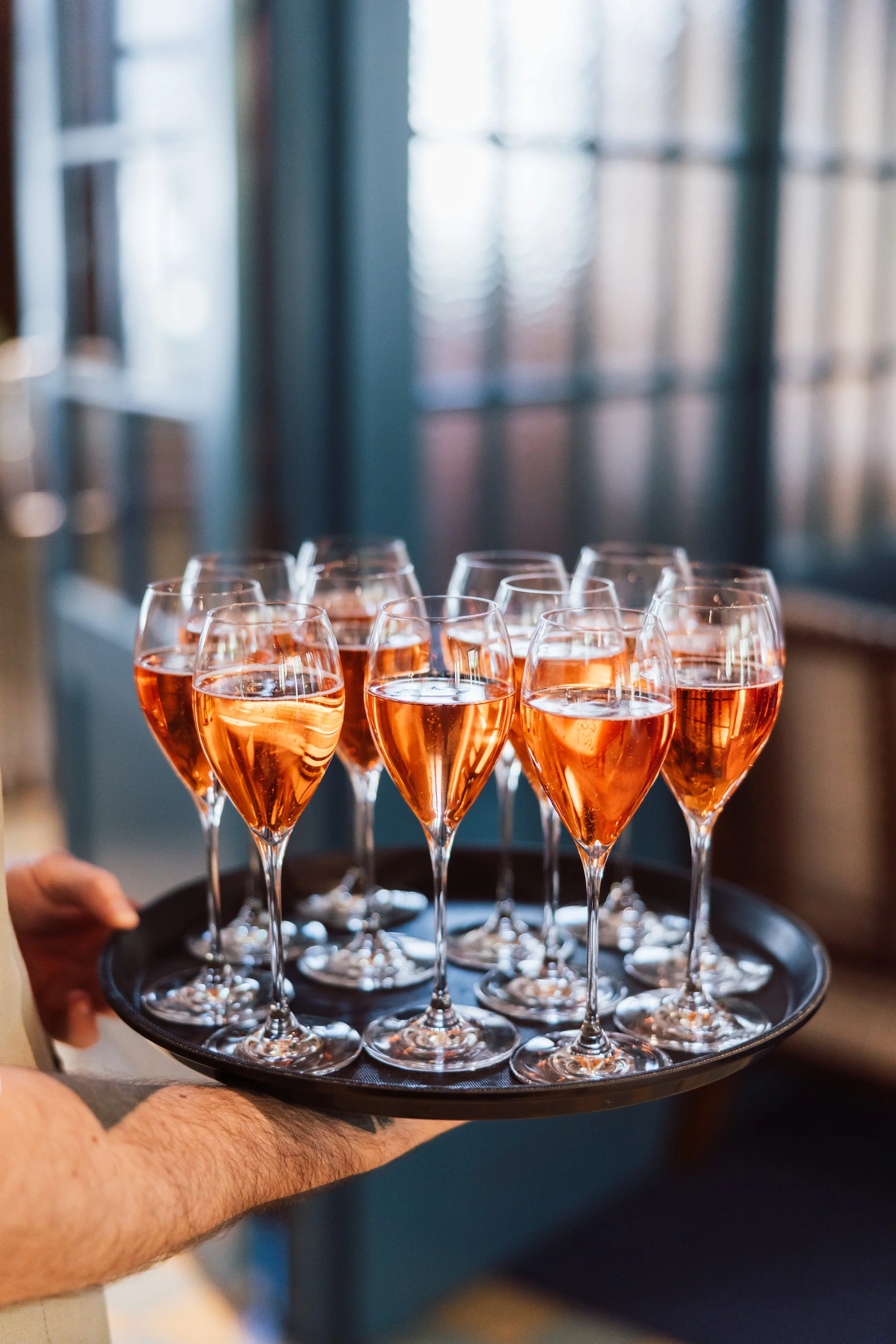 Tray of multiple glasses filled with a pink-colored beverage, being held by a person with a visible arm.