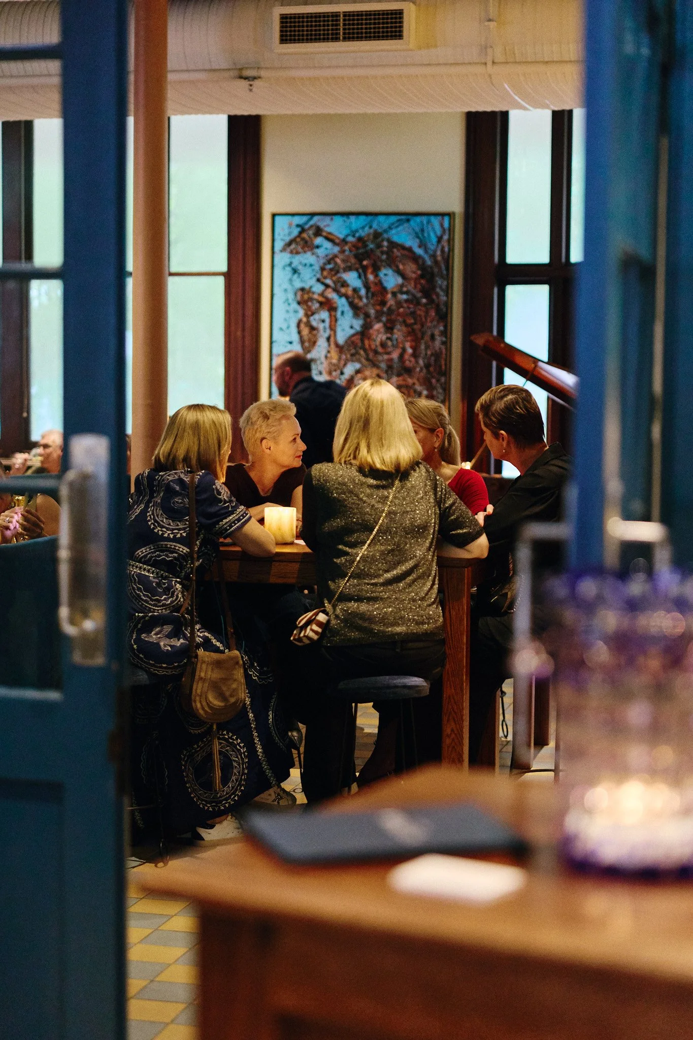 People sitting around a table engaged in conversation in a cozy indoor setting with large windows and a colorful abstract painting on the wall.