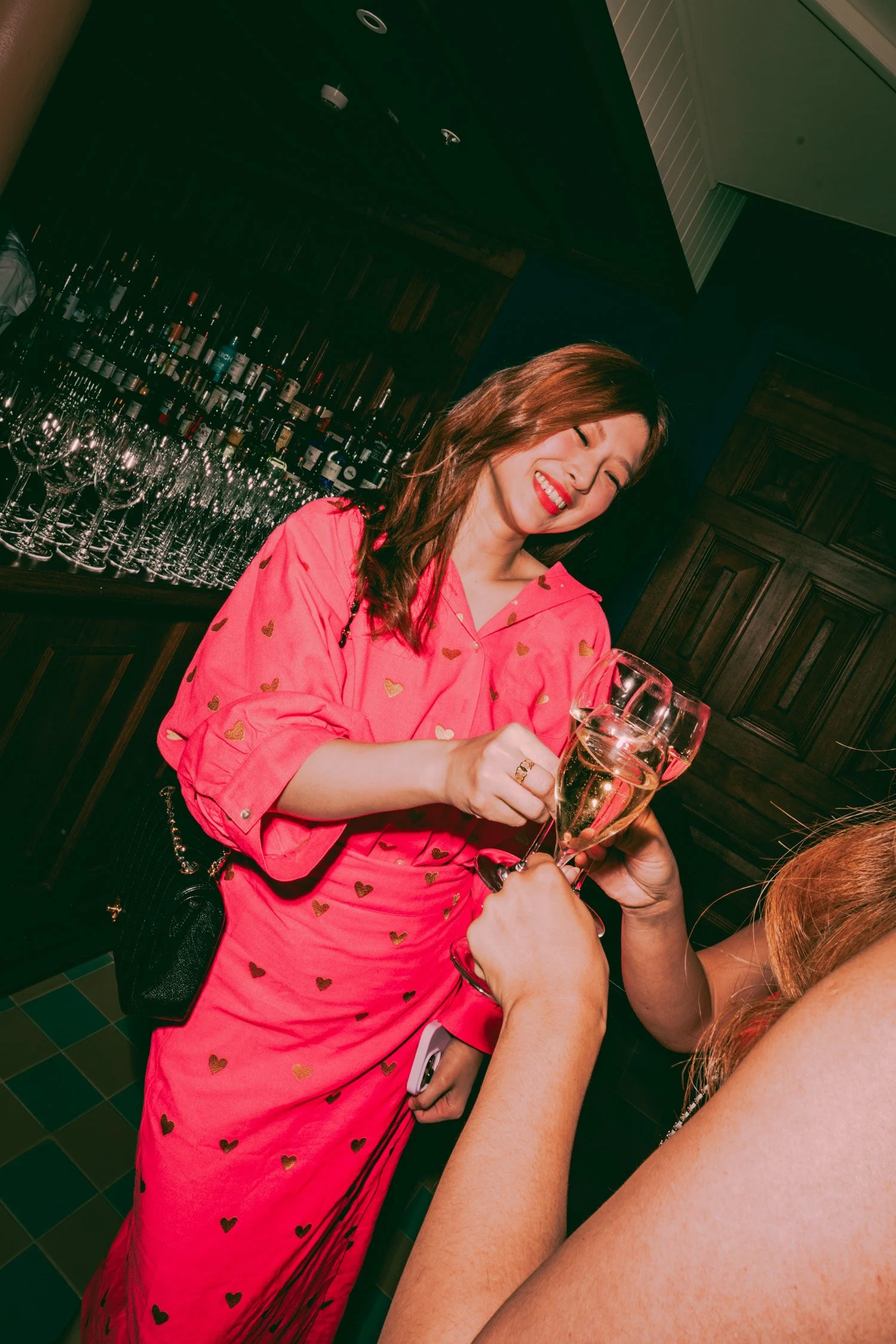 A woman in a pink dress with gold hearts smiling and holding a glass of white wine at a bar, with bottles and glasses in the background.