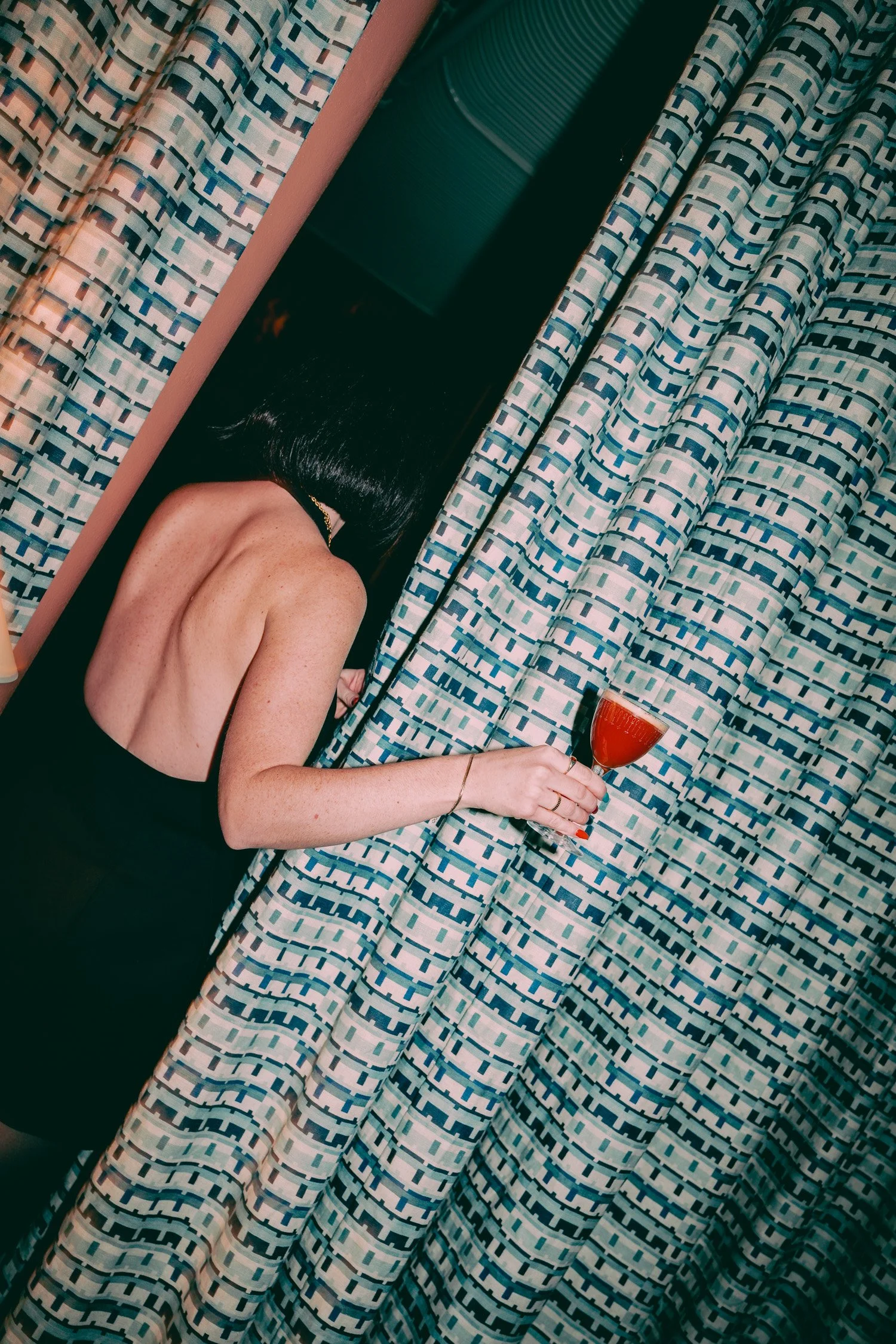 A woman with black hair in a black dress is leaning over a table, holding a glass of red drink, with patterned curtains in the background.