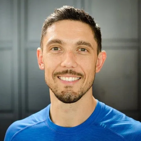 Smiling man in blue shirt with short hair and beard in front of a dark background.