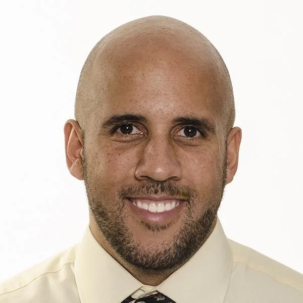 Portrait of a smiling bald man with a beard, wearing a light-colored shirt and tie.