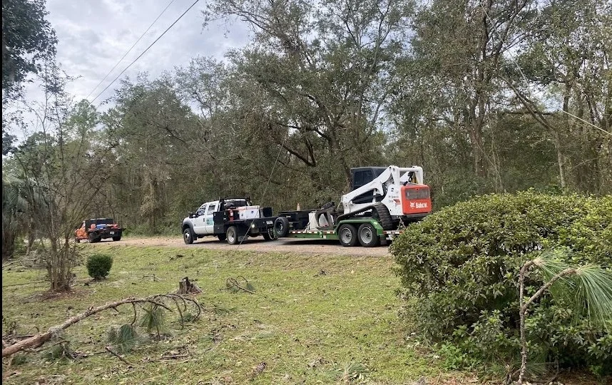 A pickup truck and a police vehicle are parked on a rural road, with a skid-steer loader on a trailer attached to the pickup truck. Tall trees and bushes surround the scene.