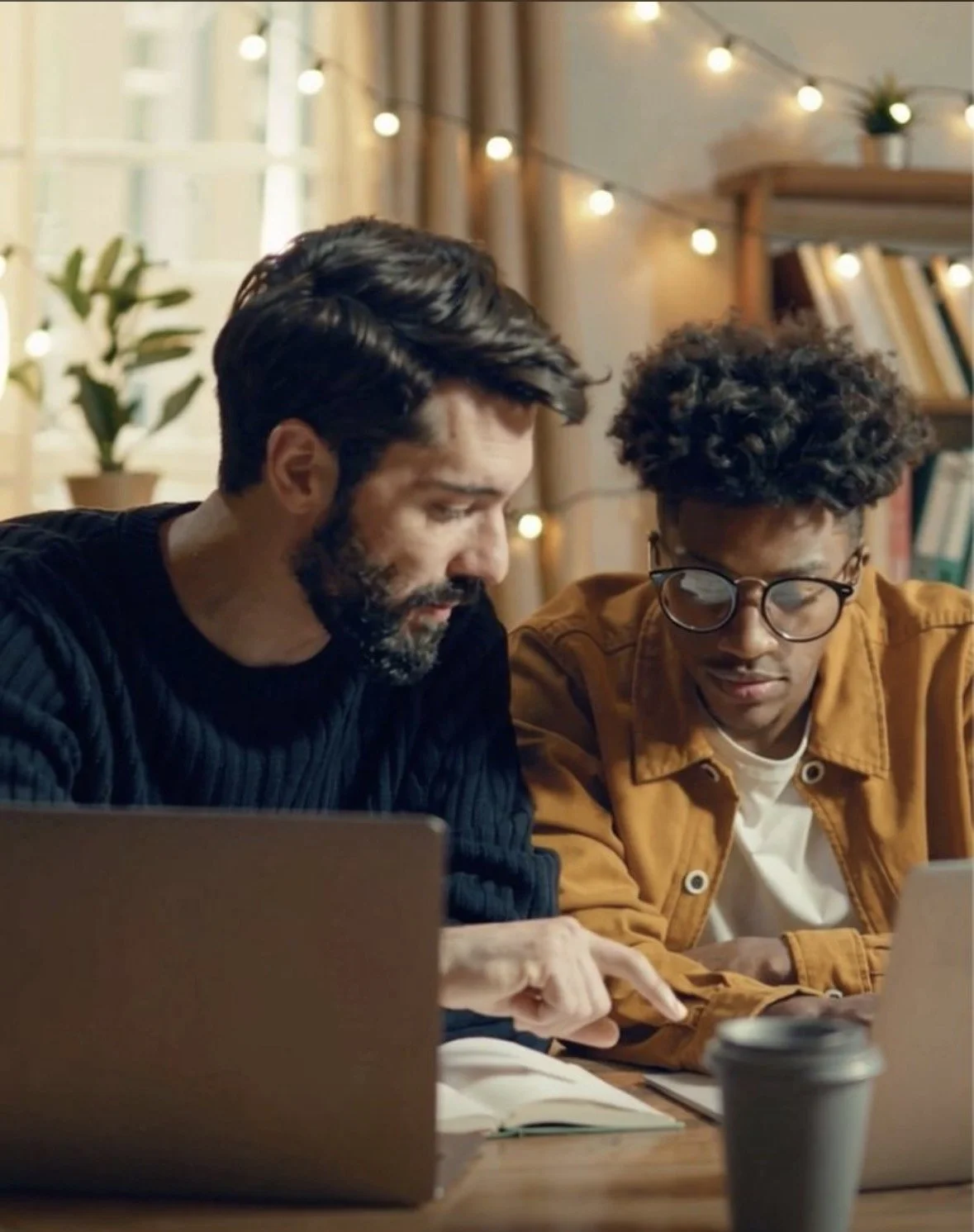 Two men, a student and tutor optimizing grades and improving education through a tutoring session looking at a laptop in a cozy room decorated with string lights and books.