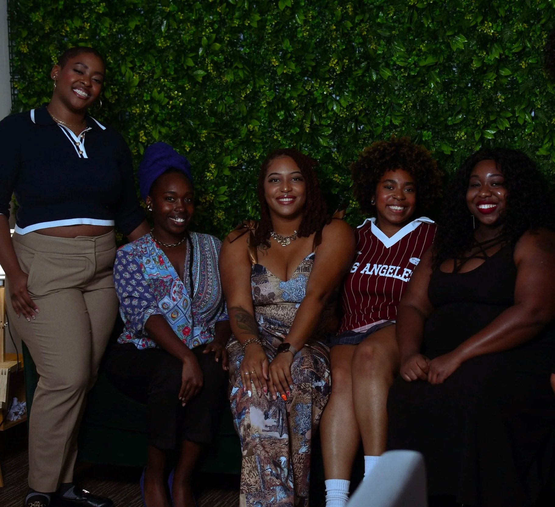 Six women sitting and standing in front of a green wall, smiling at the camera.