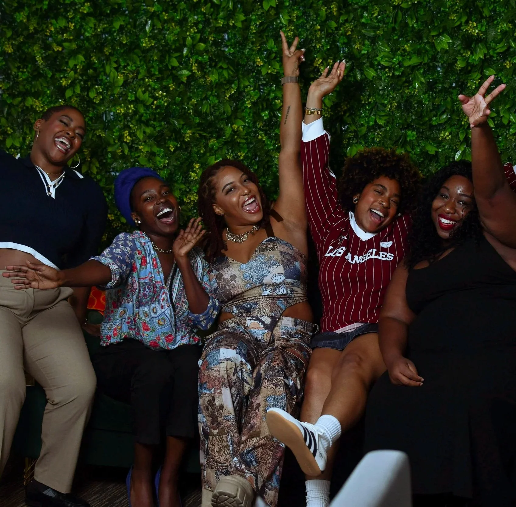Group of seven women smiling and celebrating together indoors, sitting and standing in front of a green leafy wall.